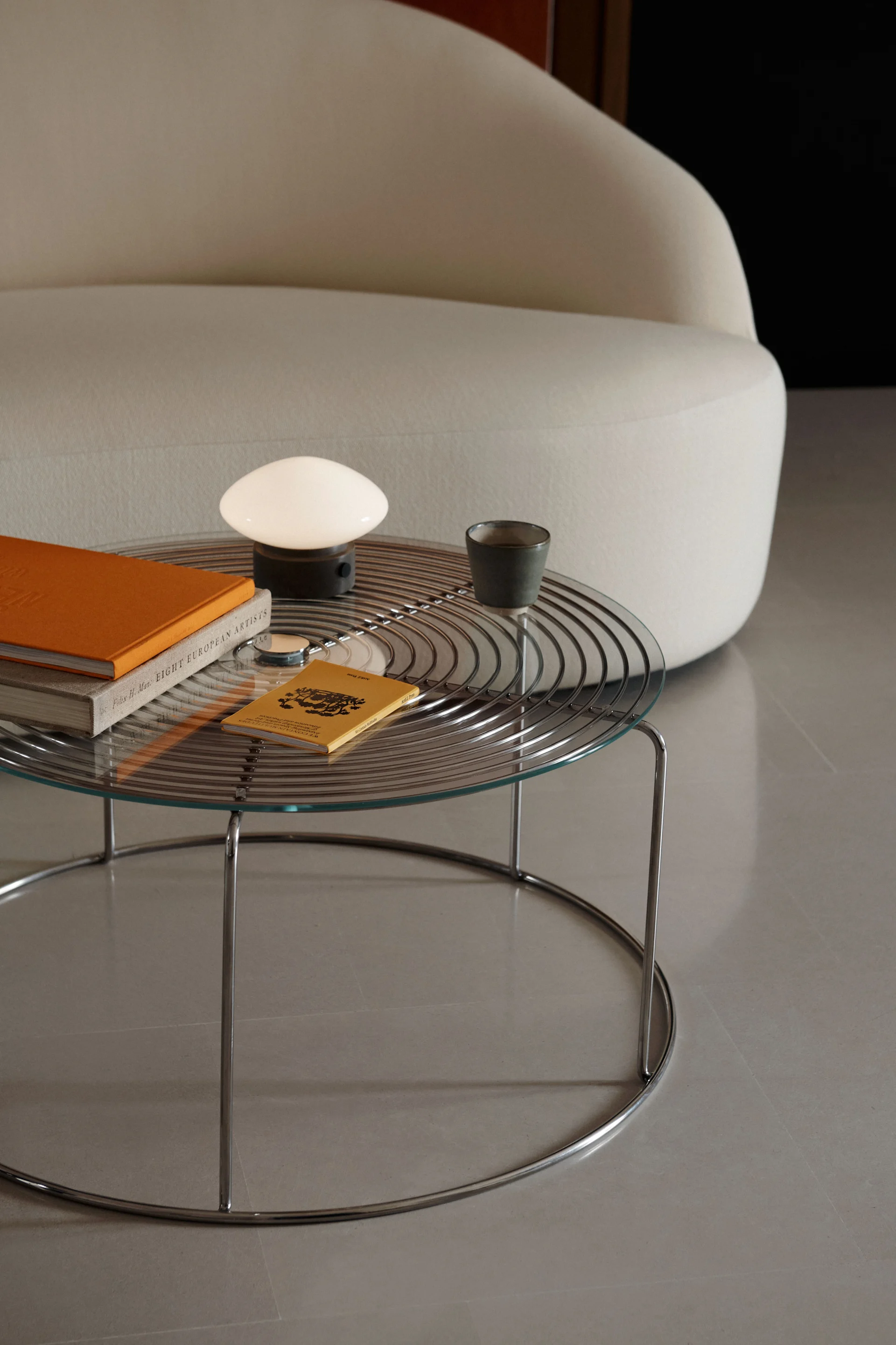 Une table basse à plateau de verre et piètement chromé supporte une lampe blanche champignon, un livre orange et une tasse sombre à côté d'un canapé crème incurvé.