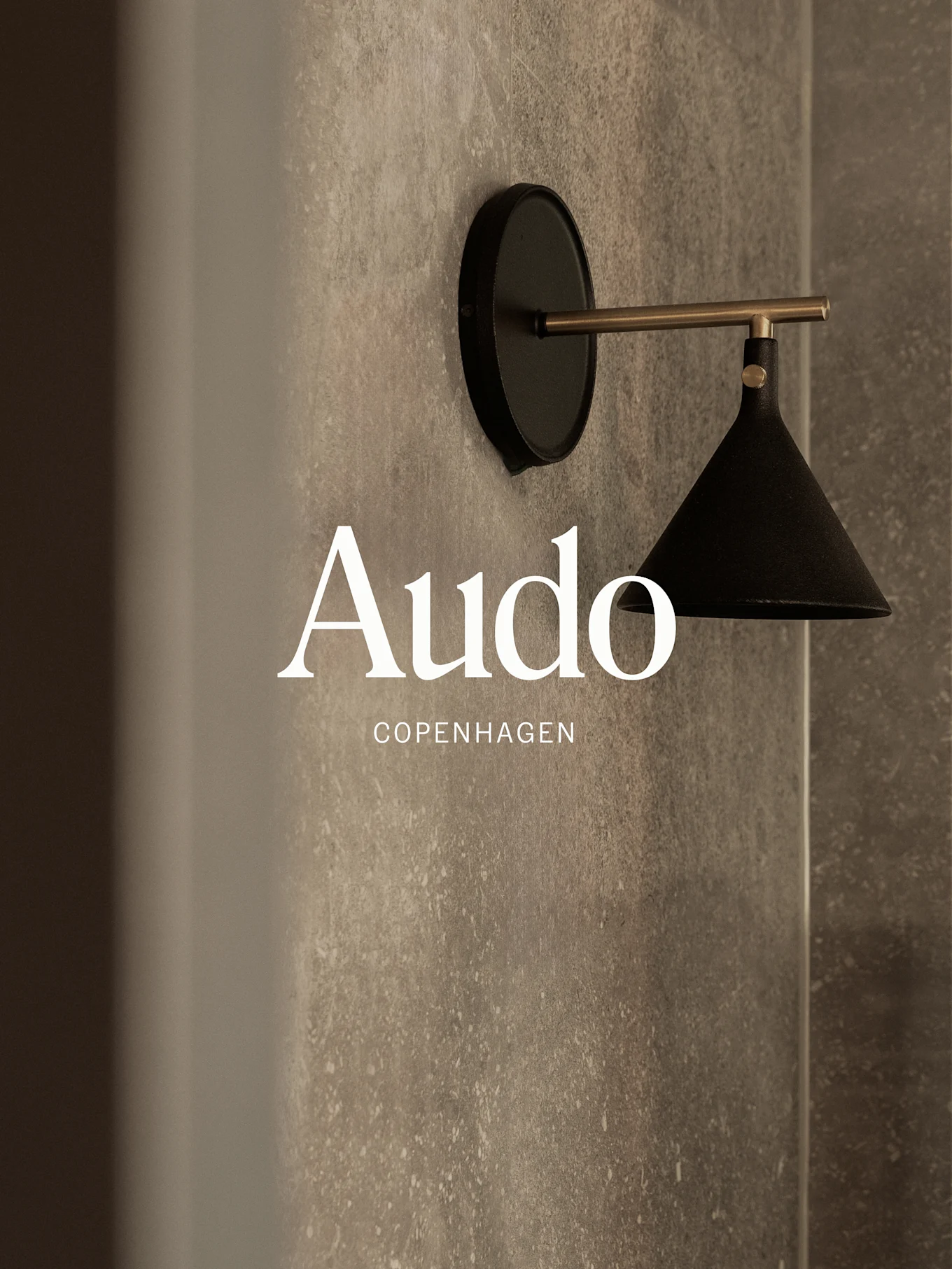 Une lampe murale noire avec un bras en laiton sur un mur gris texturé, avec le texte 'Audo Copenhagen'.