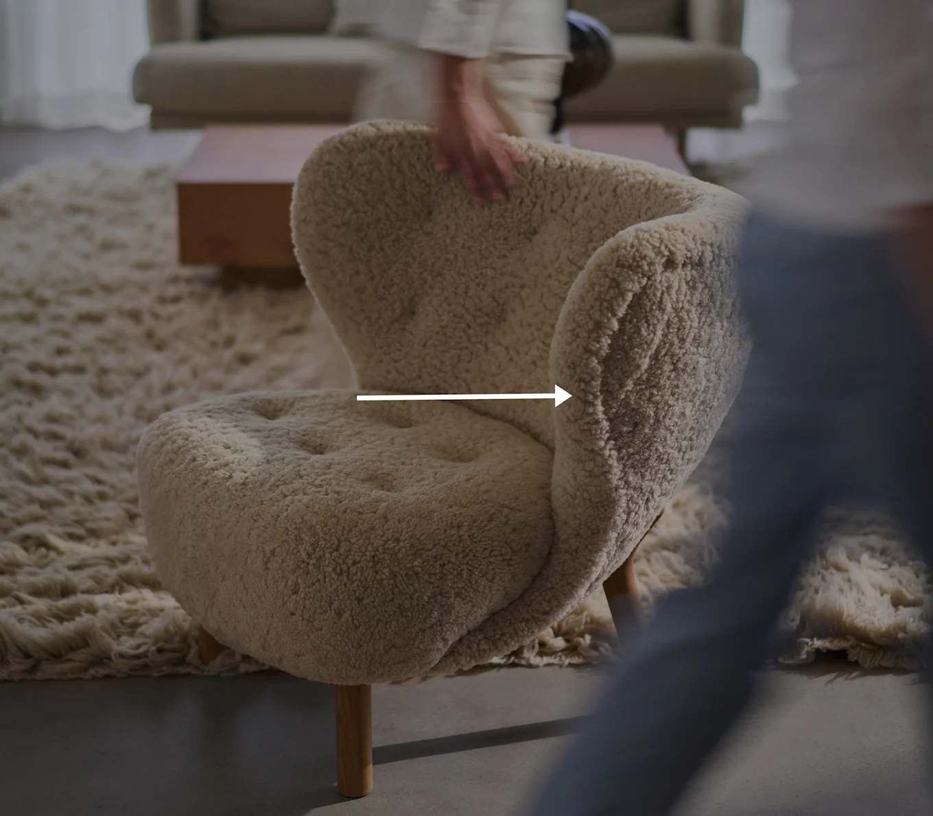 Fauteuil crème et moelleux avec pieds en bois dans un salon, une main floue touche le dossier du fauteuil.