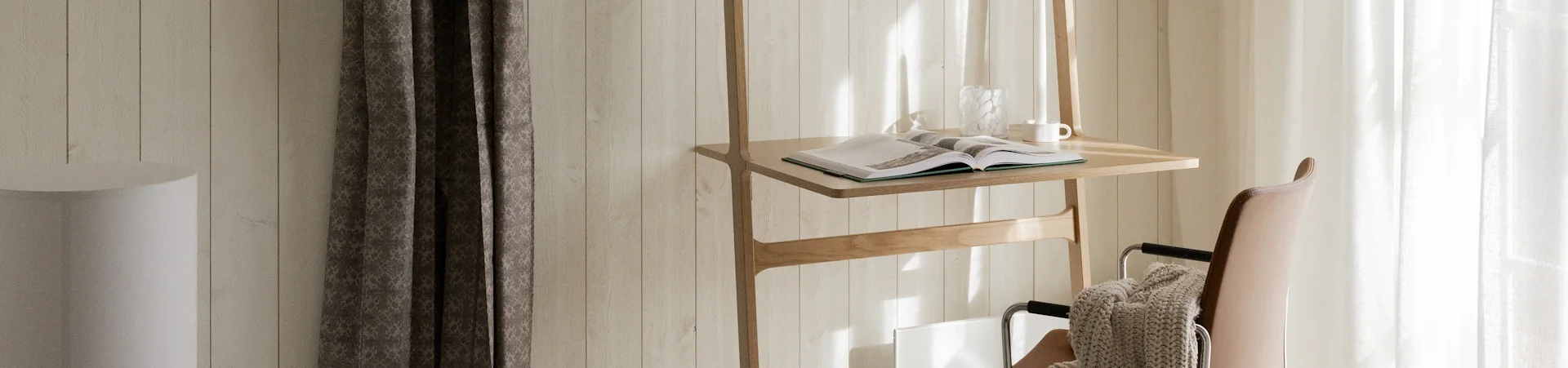 Un support en bois avec un livre ouvert, un vase en verre et une tasse blanche, à côté d'une chaise en cuir marron avec une couverture beige, baignés de soleil.