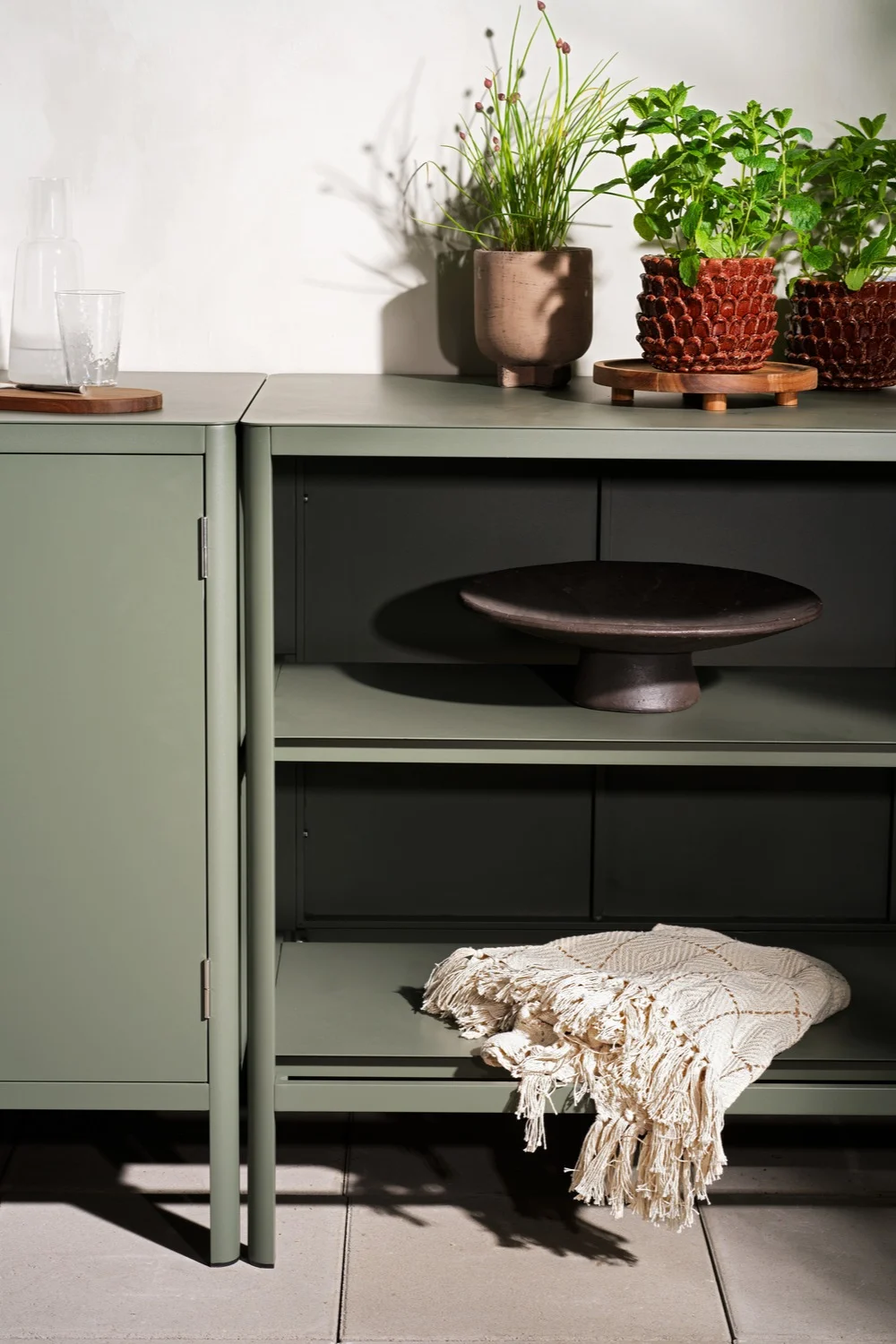 Meuble de rangement vert sauge avec des plantes en pot, une bouteille en verre, un bol décoratif et une couverture texturée.