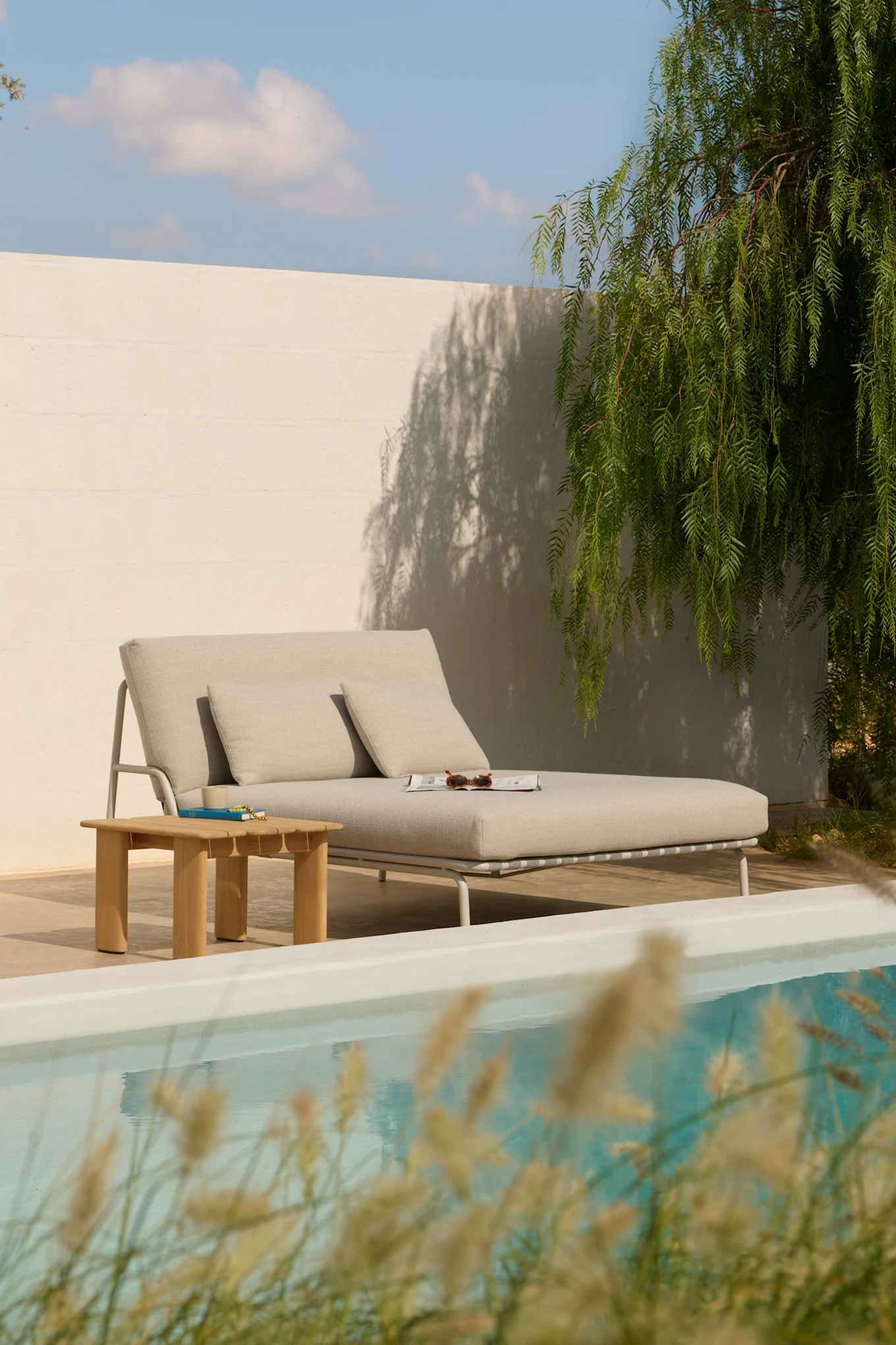 Chaiselongue extérieure gris clair avec oreillers, une table d'appoint en bois et un magazine près d'une piscine.