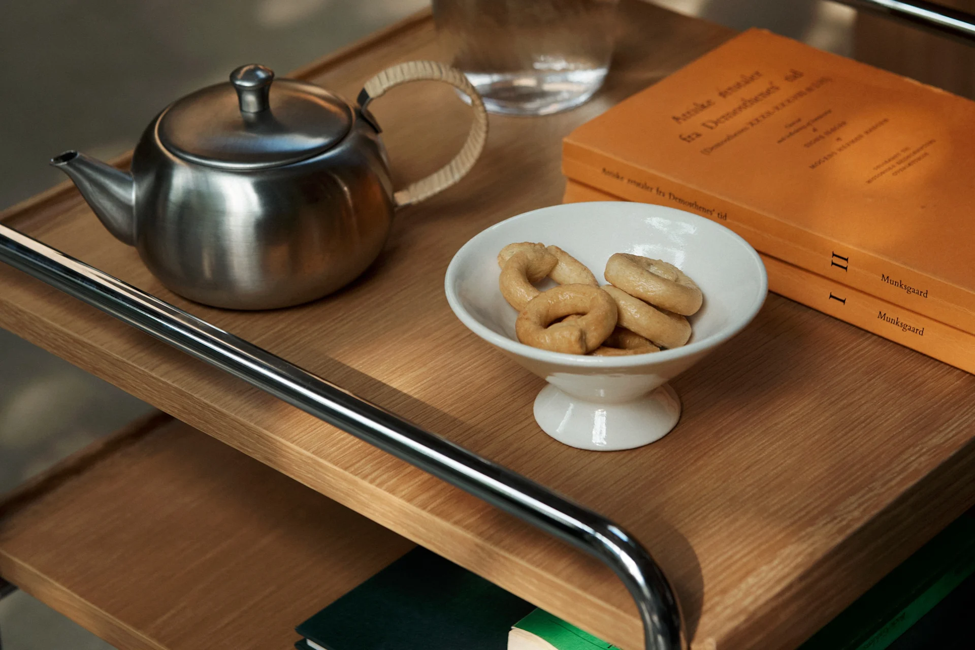 Une théière en acier inoxydable, un bol blanc avec des biscuits en forme d'anneau et deux livres orange sur une étagère en bois.
