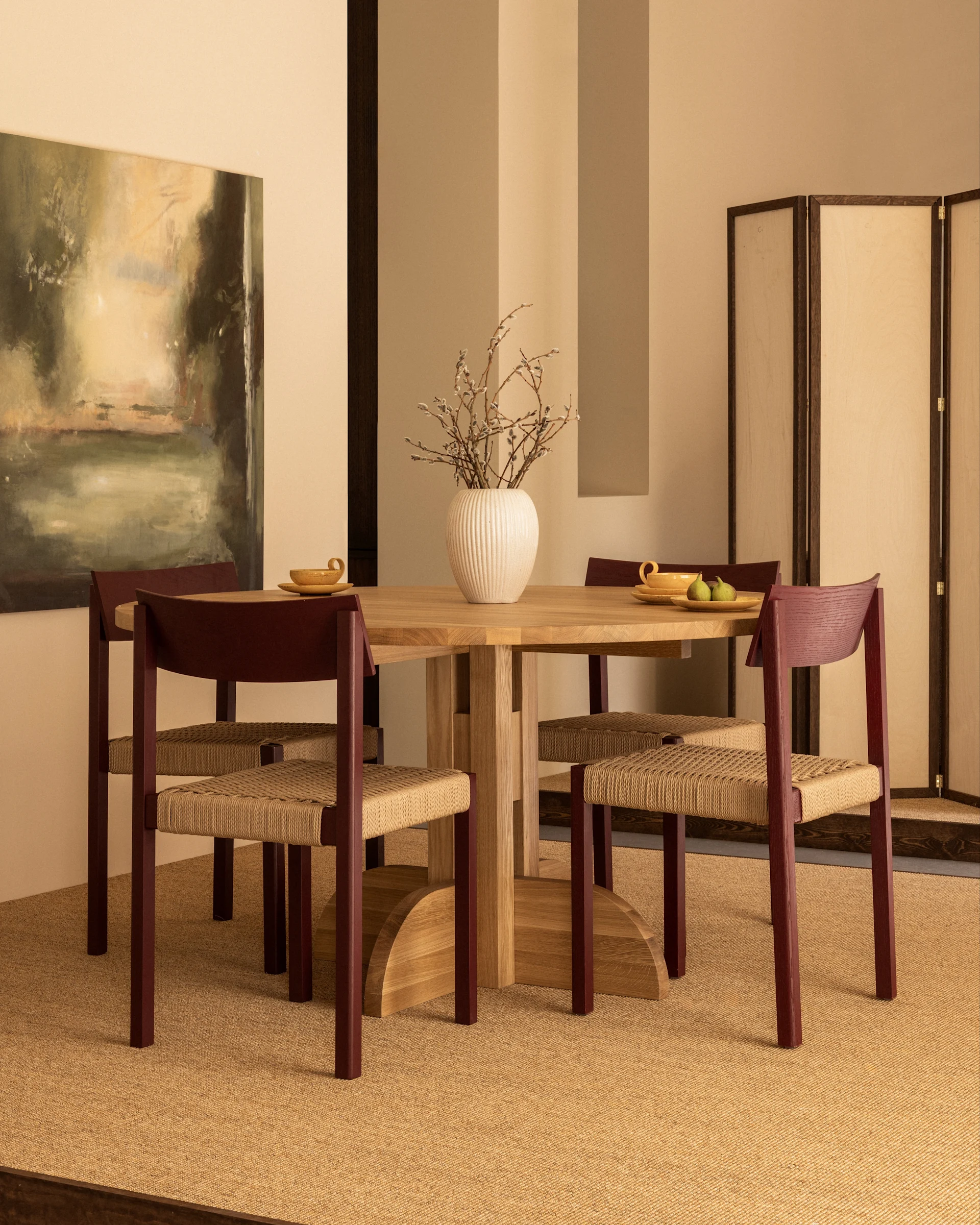 Une salle à manger avec une table ronde en bois, quatre chaises aux cadres brun rougeâtre et assises tressées, et un vase de chatons de saule.