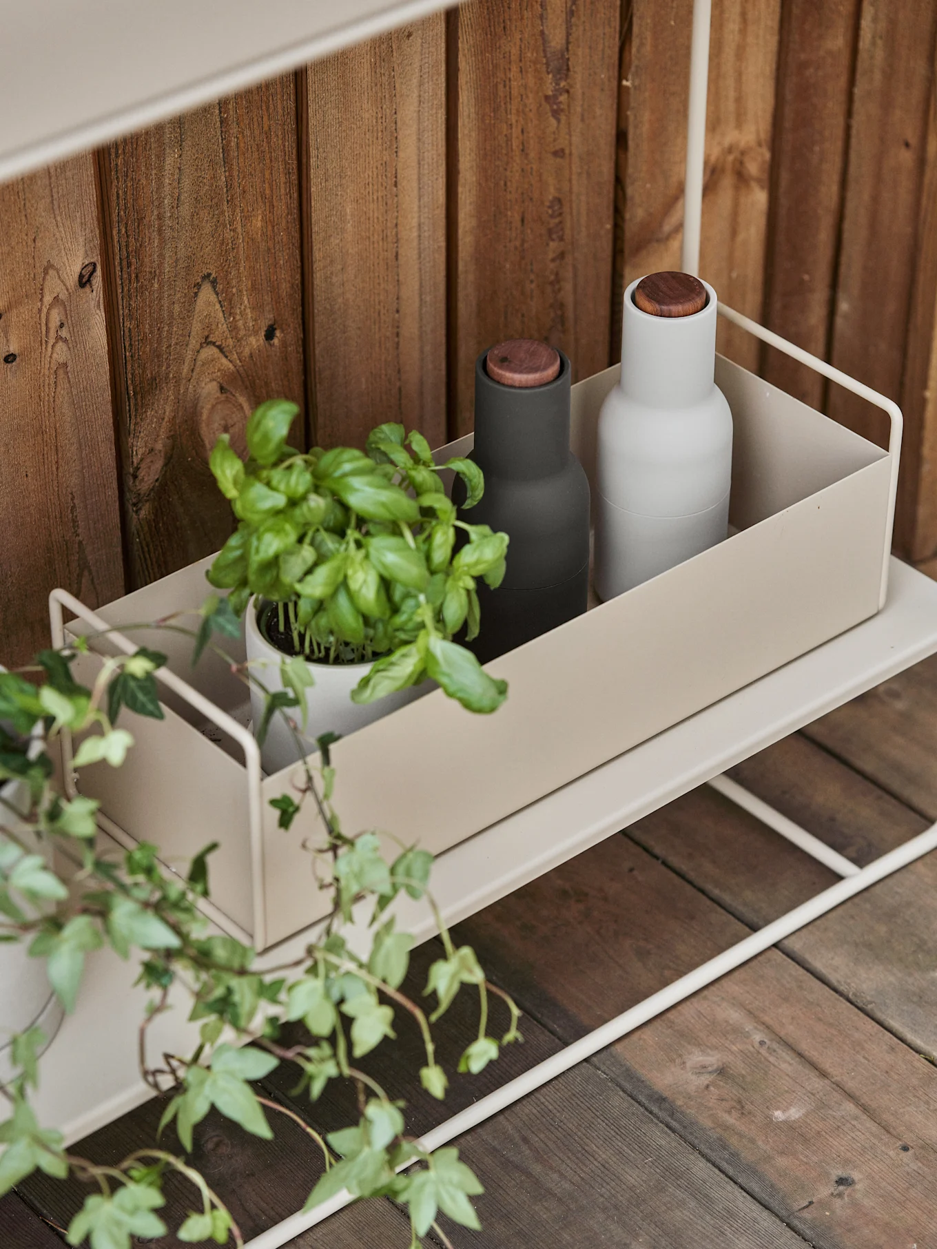 Une étagère en métal beige clair avec un récipient contenant un plant de basilic, un moulin noir et un moulin blanc sur fond de bois.