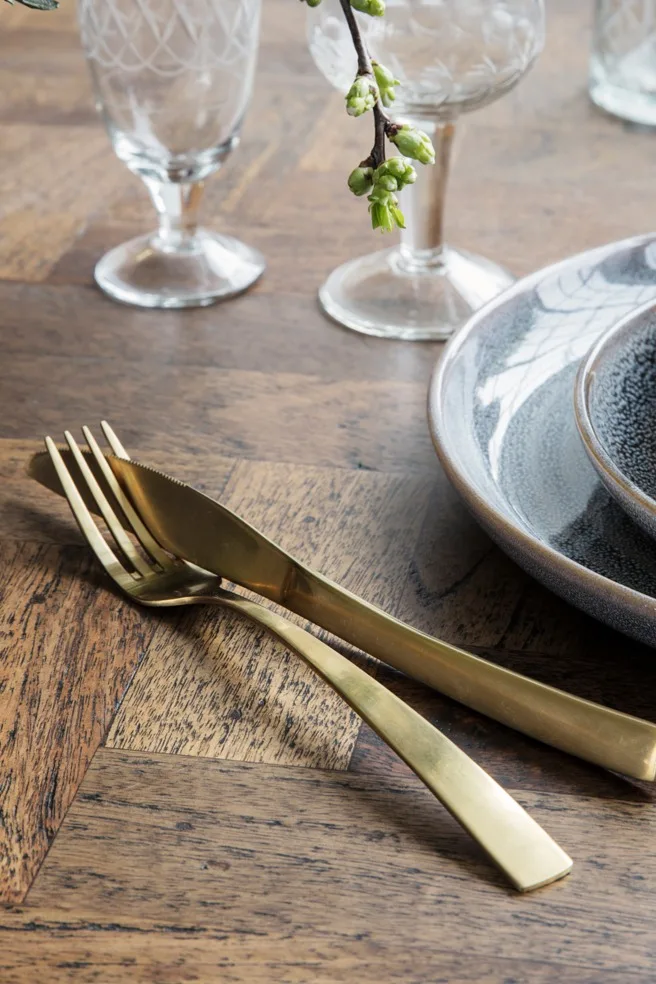 Fourchette et couteau dorés sur une table en bois à côté d'assiettes en céramique et de verres à motifs, avec une petite branche de bourgeons verts.