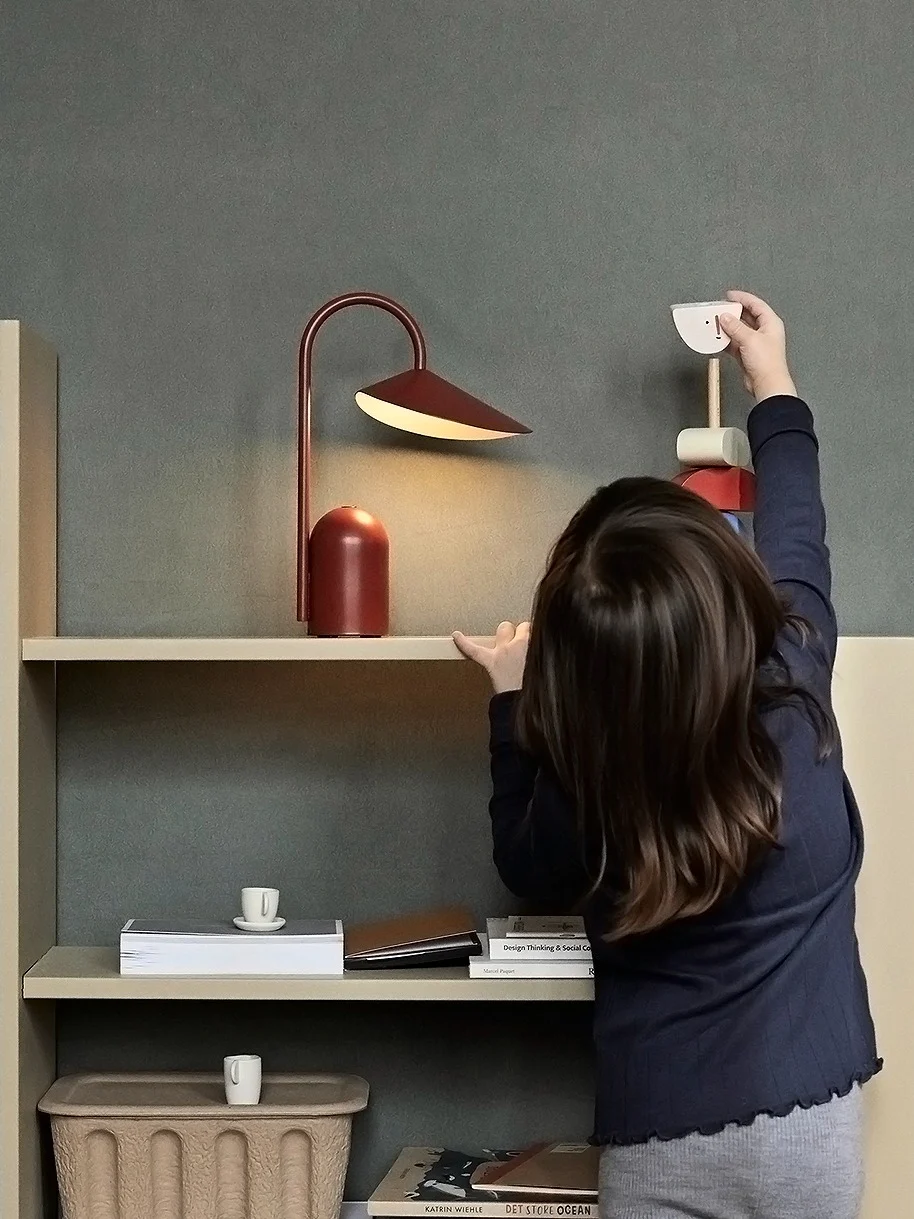 Un enfant attrape un jouet oiseau en bois sur une étagère claire avec une lampe rouge et des livres; un panier tressé est au sol.