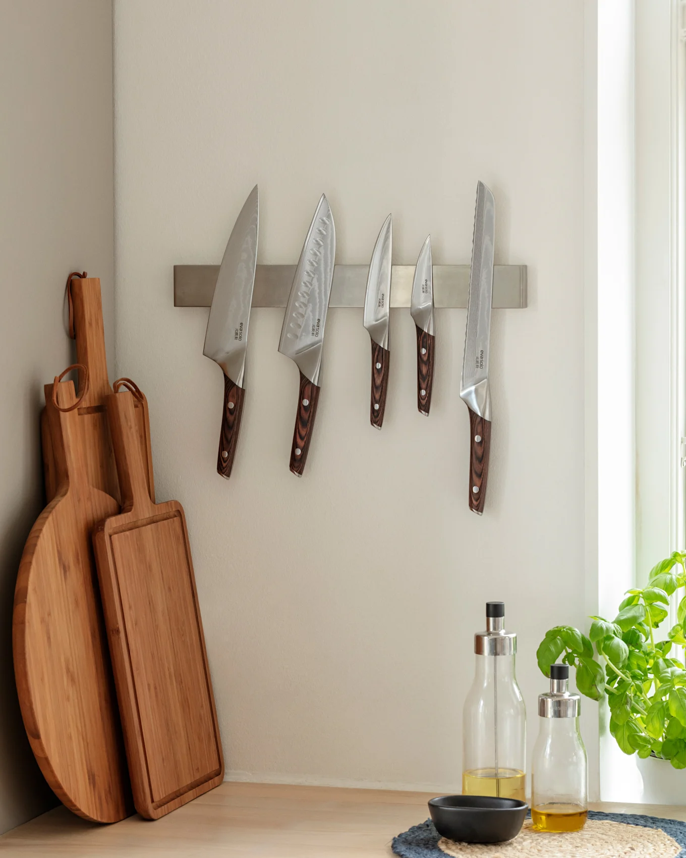 Coin de cuisine avec cinq couteaux sur une bande magnétique, deux planches à découper en bois et deux bouteilles d'huile d'olive sur un comptoir en bois à côté d'un plant de basilic.