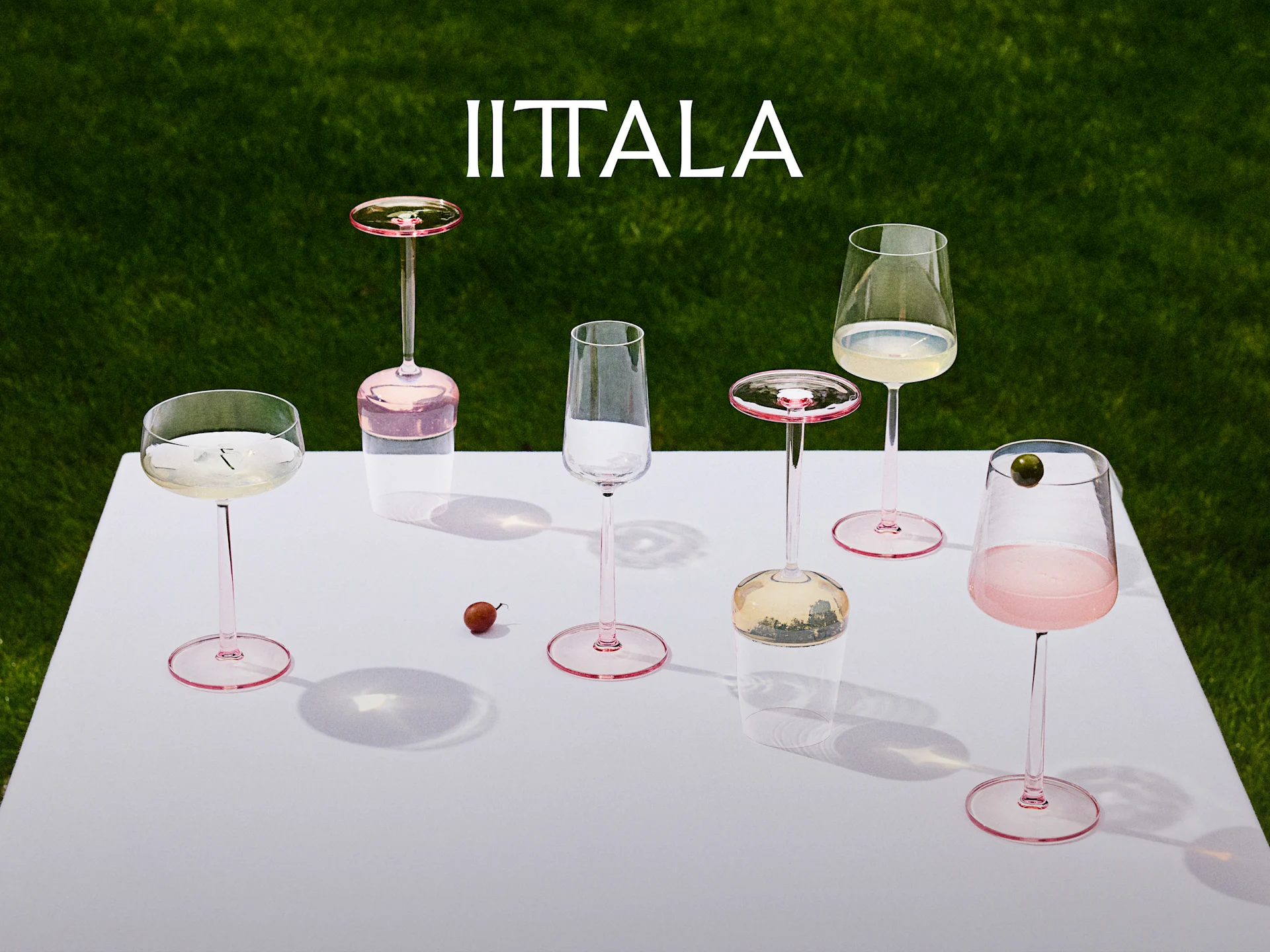 Logo Iittala au-dessus d'une collection de verres à pied roses sur une table blanche avec un fond d'herbe verte, certains avec des boissons.