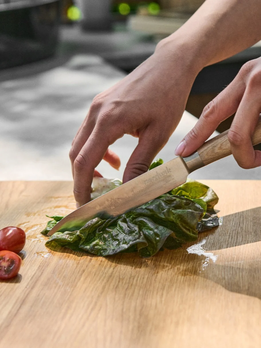 Mains coupant de la laitue verte fraîche sur une planche à découper en bois avec un couteau, à côté de tomates cerises coupées.