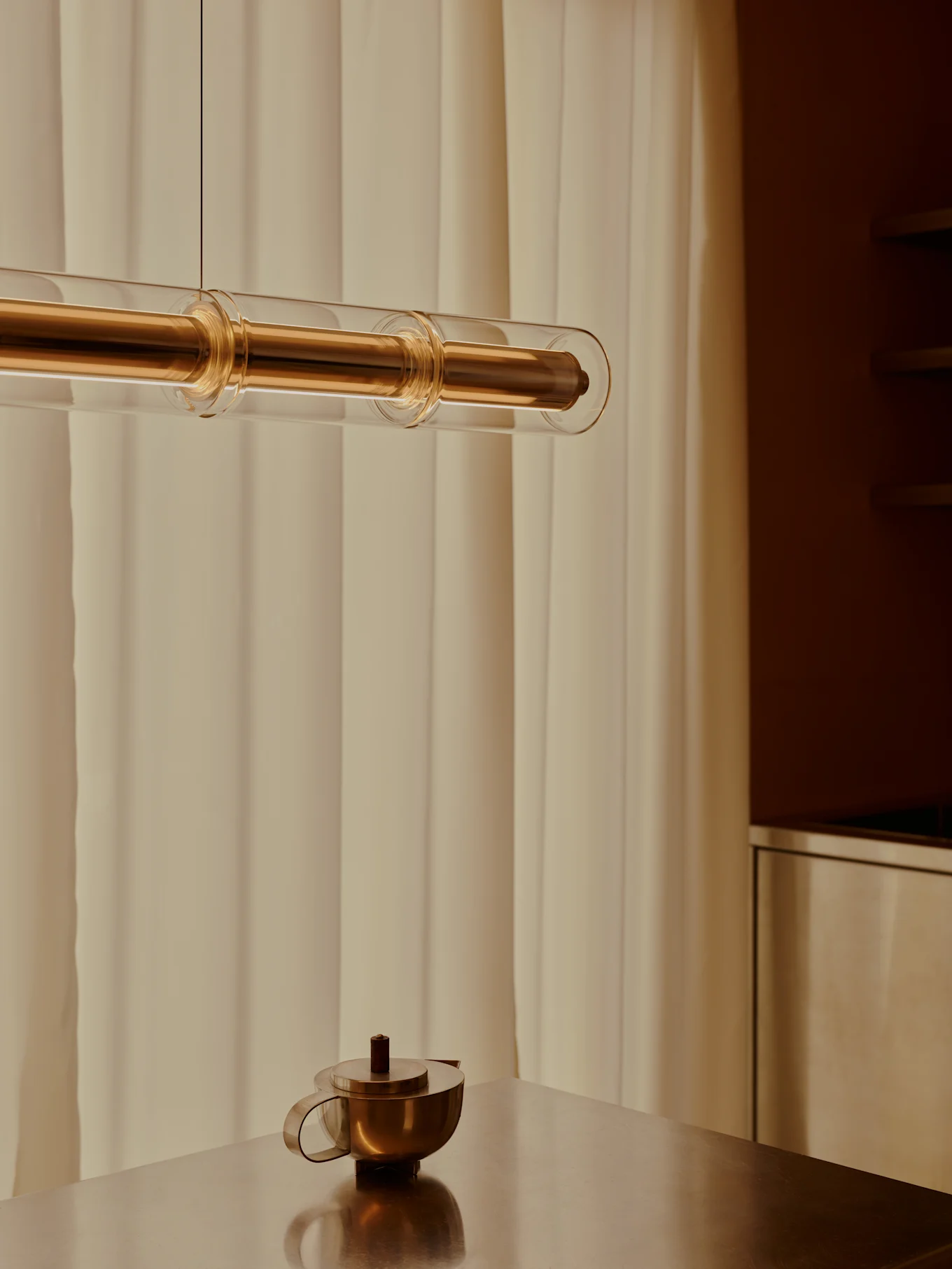 Une suspension moderne en laiton et tubes de verre est suspendue au-dessus d'une théière en laiton sur une table métallique, avec des rideaux clairs en arrière-plan.