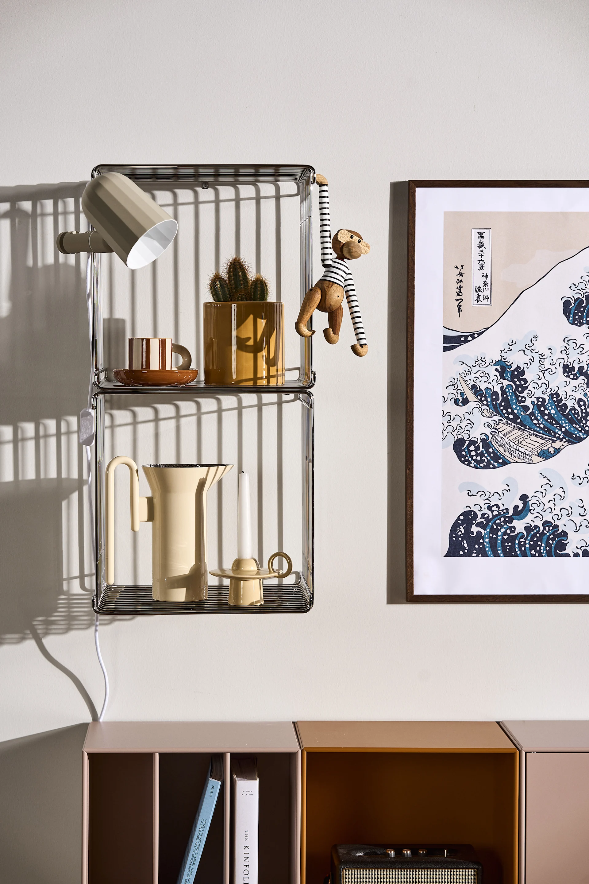 Deux étagères en treillis métallique superposées, avec une applique murale, une tasse rayée, un cactus en pot, un jouet en forme de singe, une théière et une bougie, à côté d'une estampe japonaise représentant des vagues.