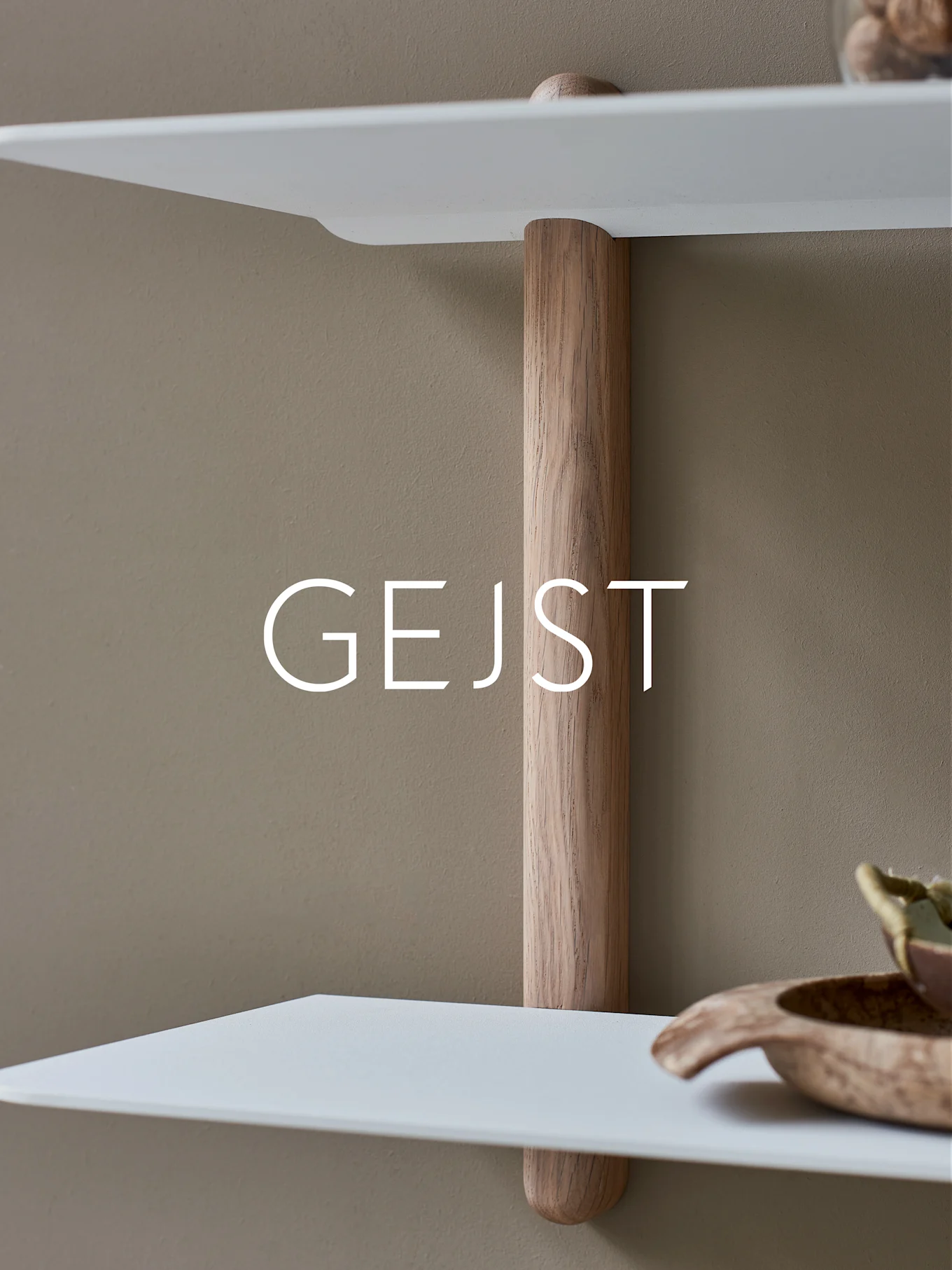 Deux étagères blanches et arrondies reliées par un poteau de support vertical en bois clair sur un mur beige. Un bol en bois est sur l'étagère inférieure.
