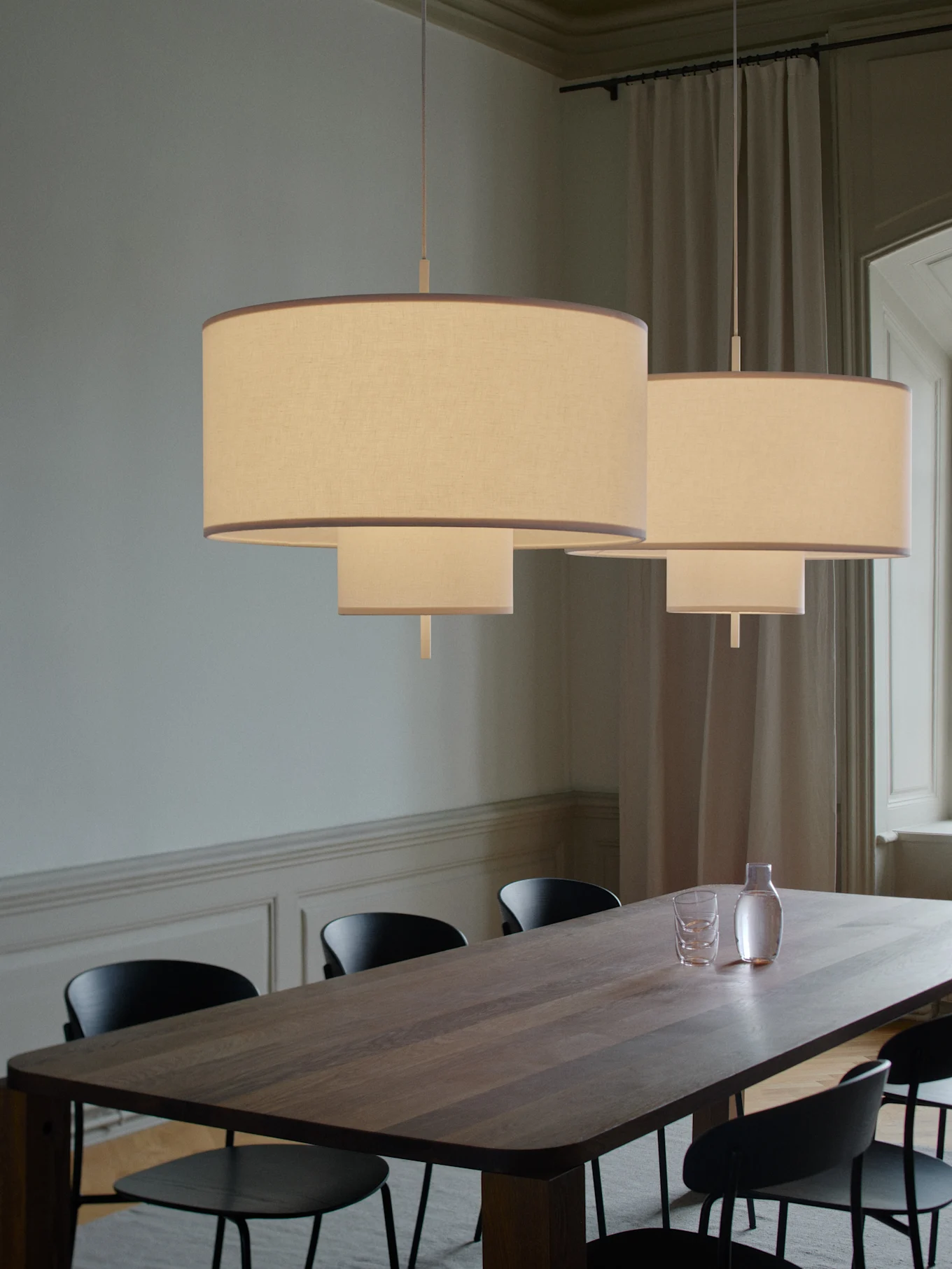 Deux suspensions en tissu étagées éclairent une longue table à manger en bois avec des chaises noires, une carafe et des verres.