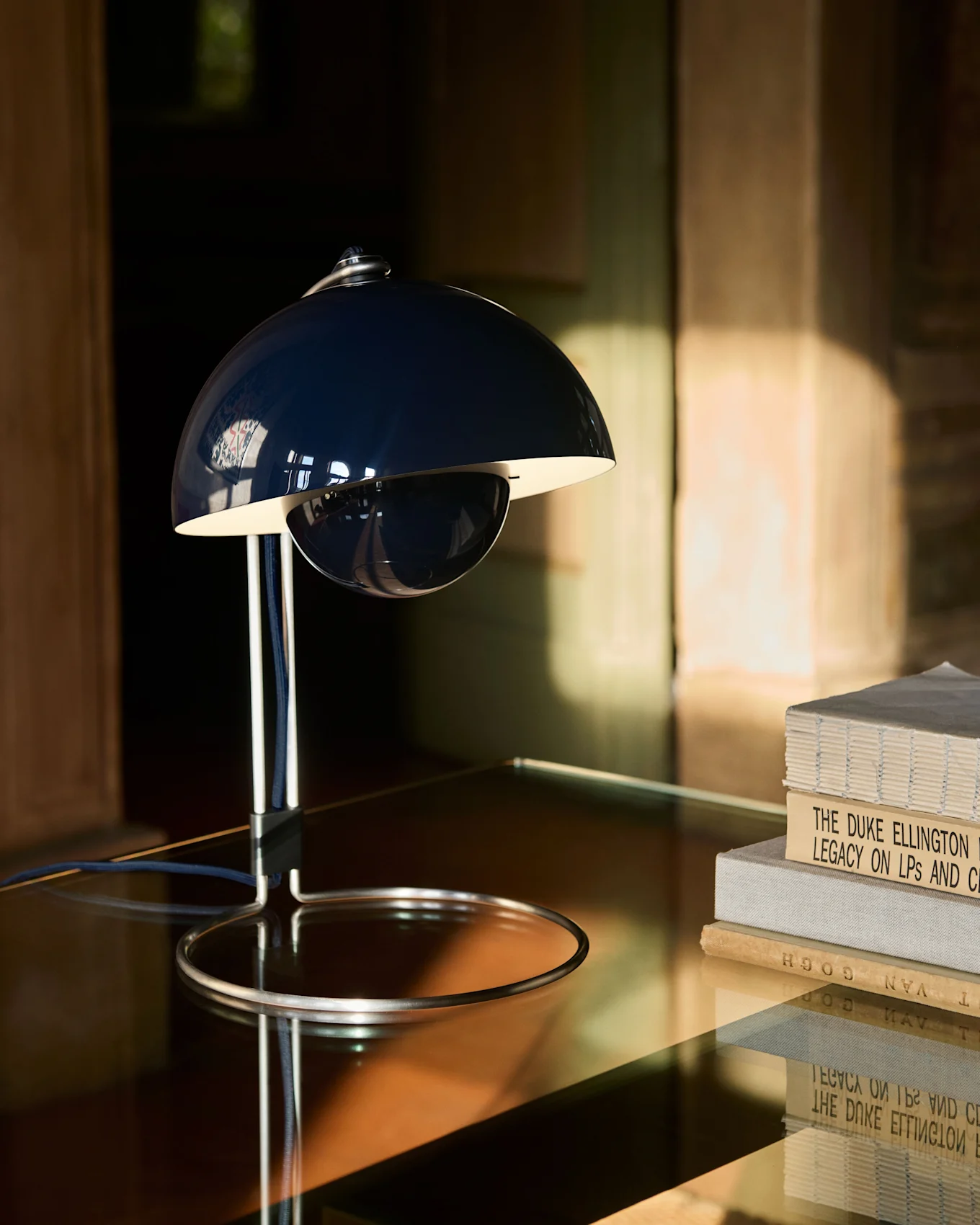 Une lampe de table bleu foncé en forme de cloche avec une base chromée sur une table réfléchissante, à côté d'une pile de livres.
