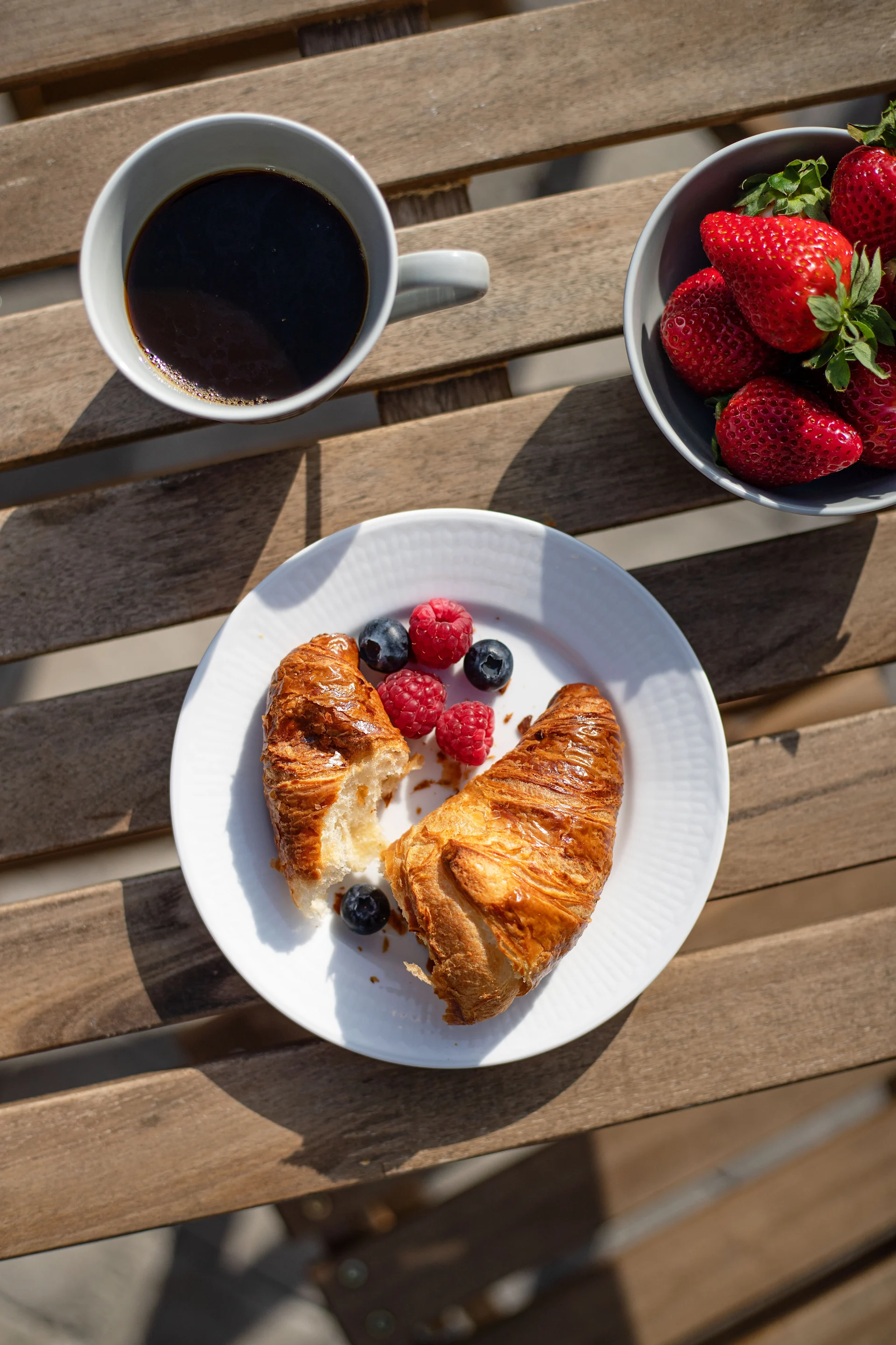 Vue plongeante d'un petit-déjeuner sur une table en bois avec café, fraises, croissants, myrtilles et framboises.