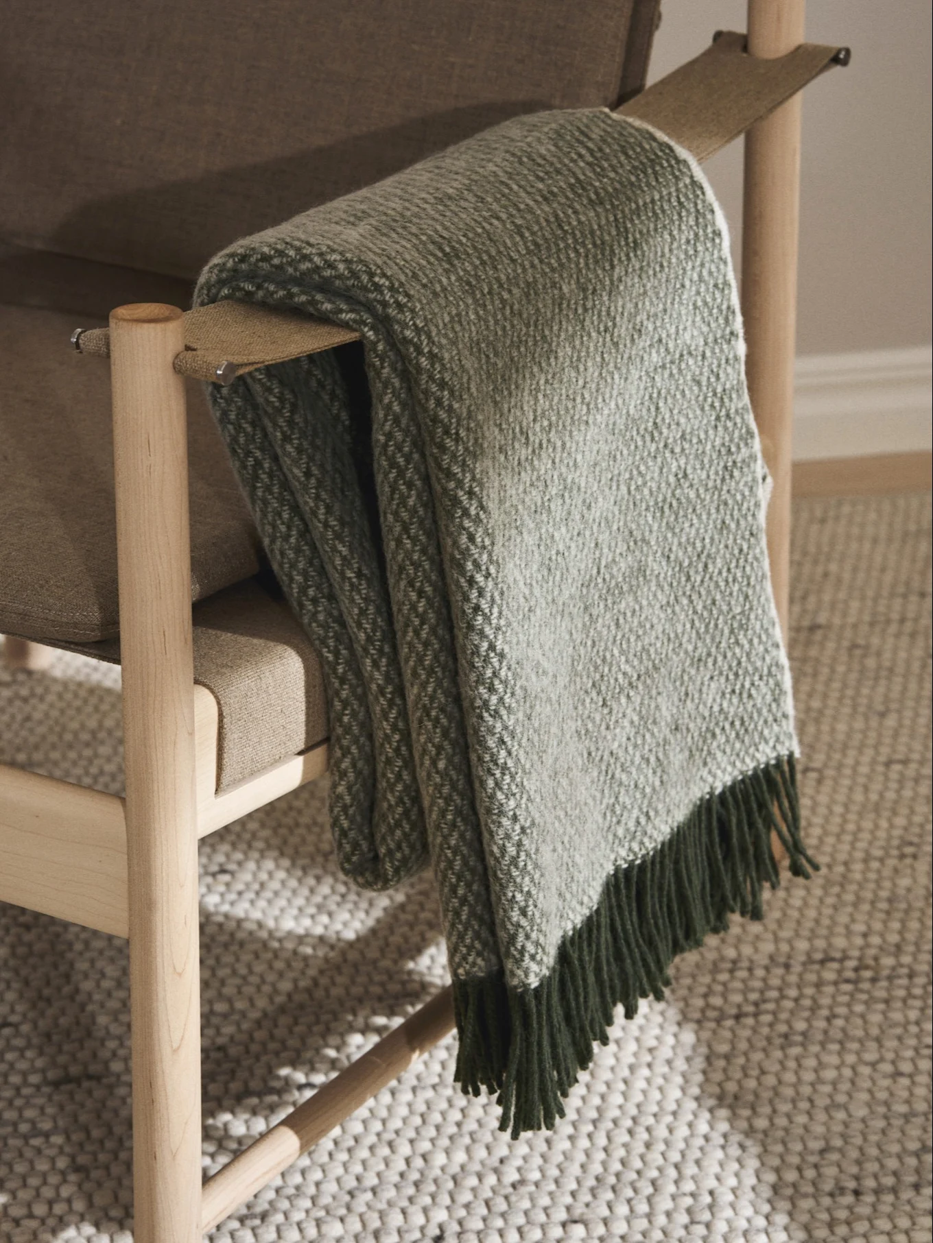 Un fauteuil en bois marron clair avec une couverture à motifs verts et blancs drapée sur l'accoudoir, avec des franges vert foncé.