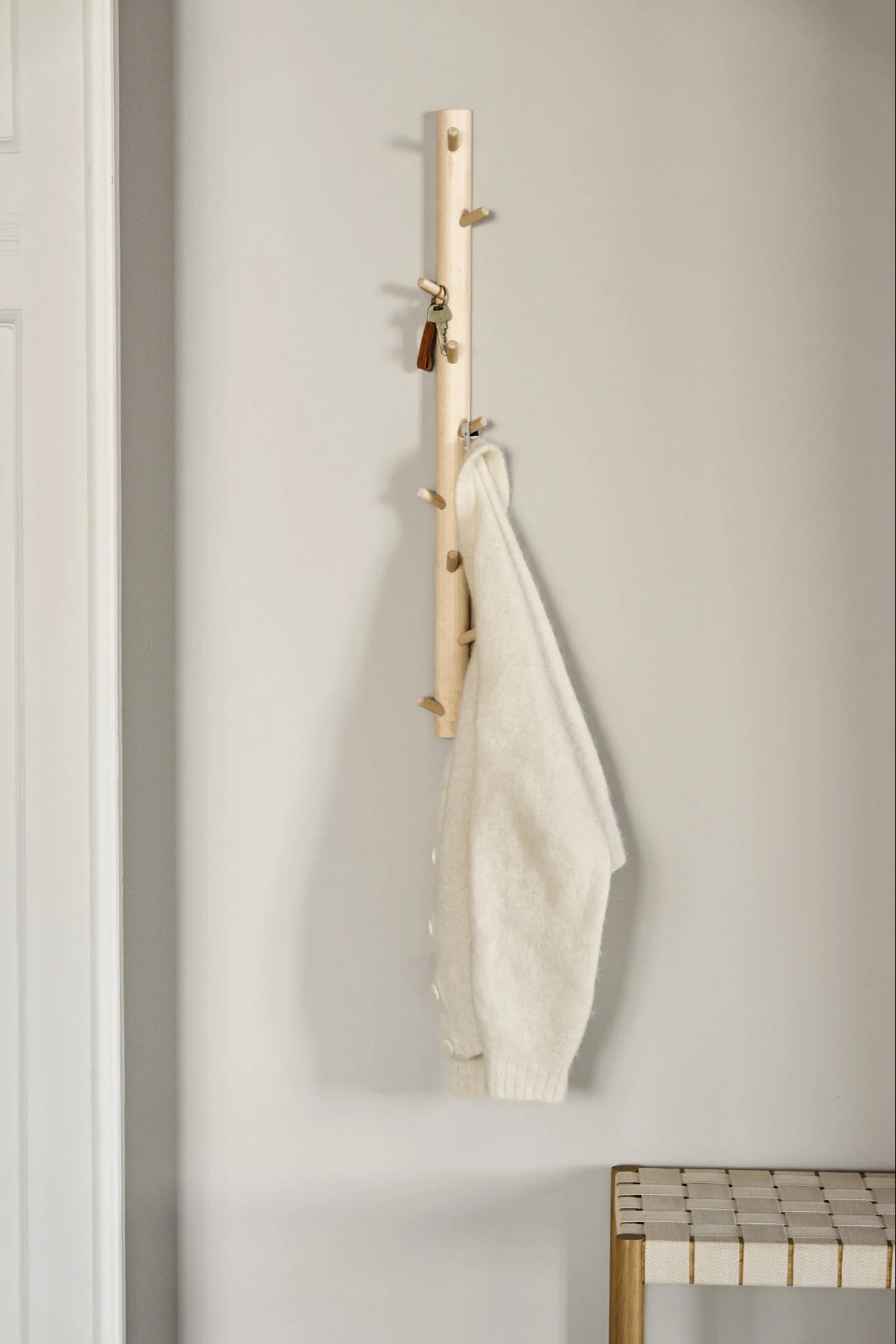 Un porte-manteau en bois clair avec des patères tient des clés et un pull crème, avec un banc tressé en dessous.