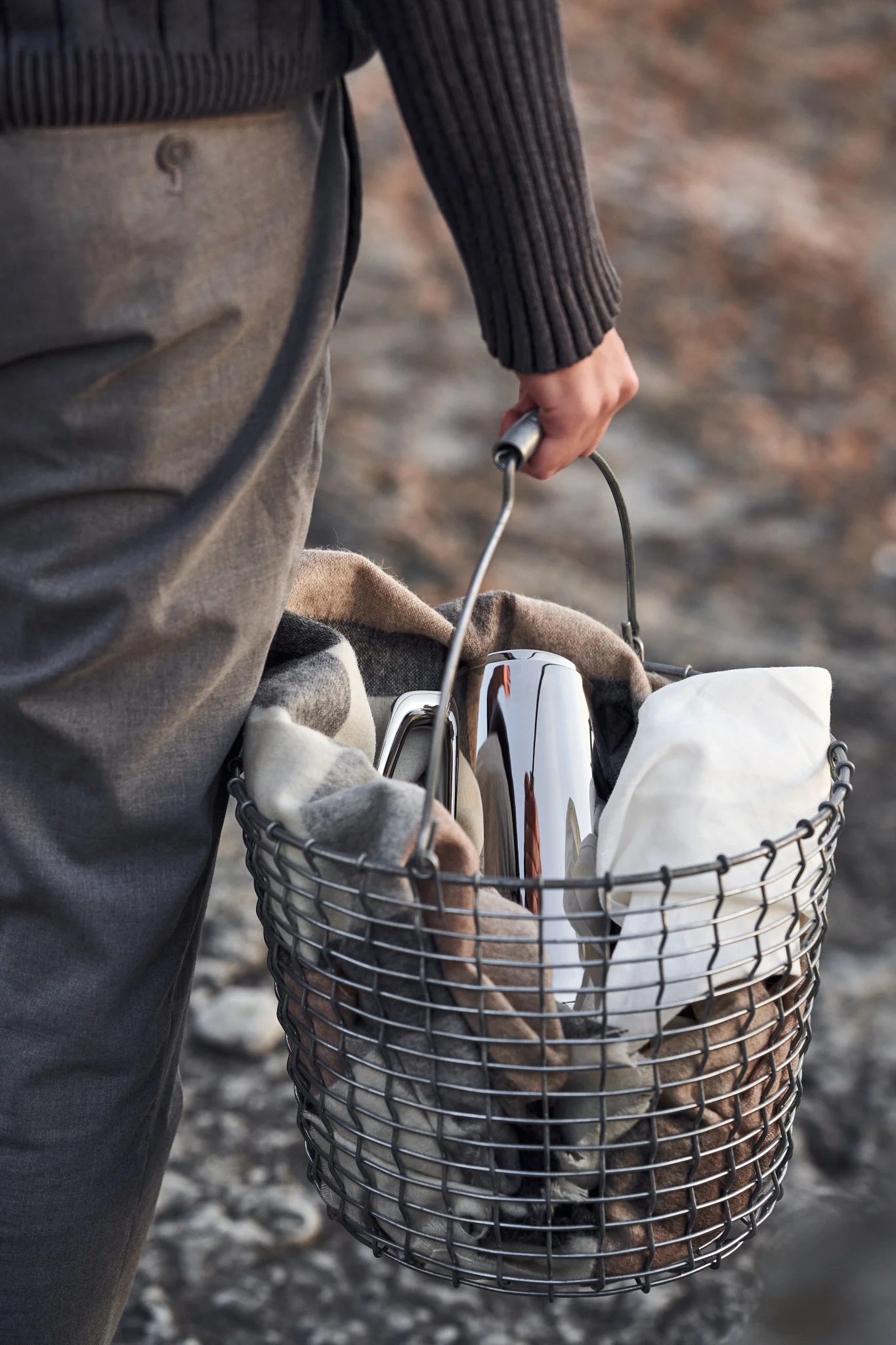 Une personne porte un panier en métal contenant une couverture à carreaux, un thermos argenté et un linge blanc.