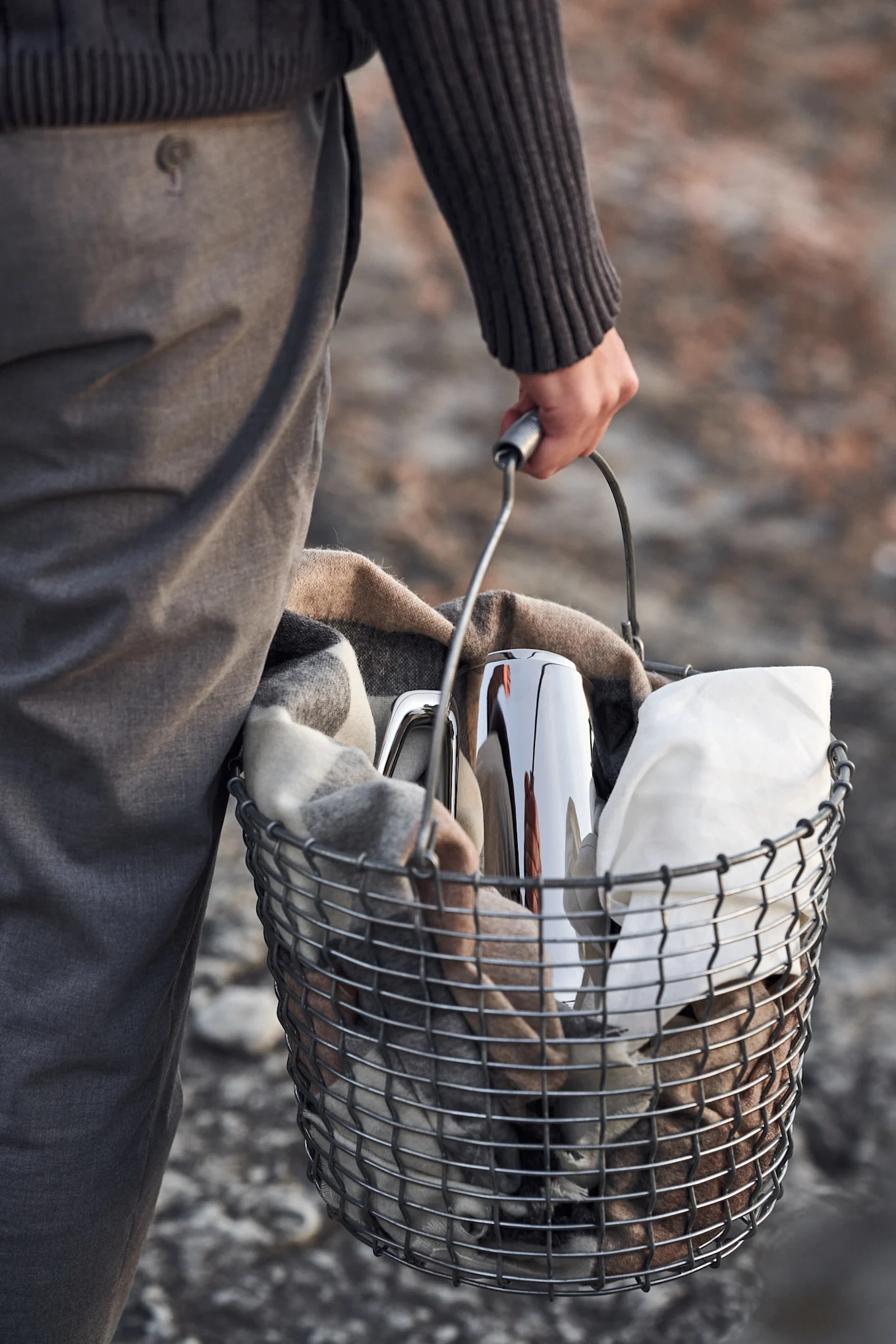 Une personne porte un panier en métal contenant une couverture à carreaux, un thermos argenté et un linge blanc.