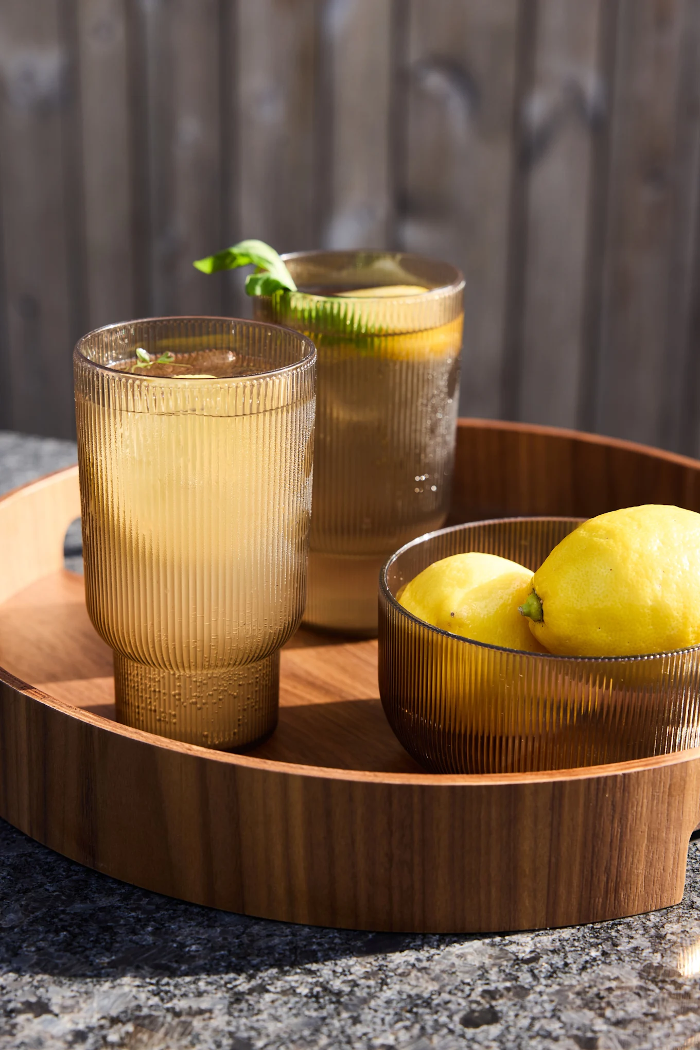 Deux verres cannelés de couleur brun doré remplis de limonade et une coupe de citrons posés sur un plateau en bois à l'extérieur.