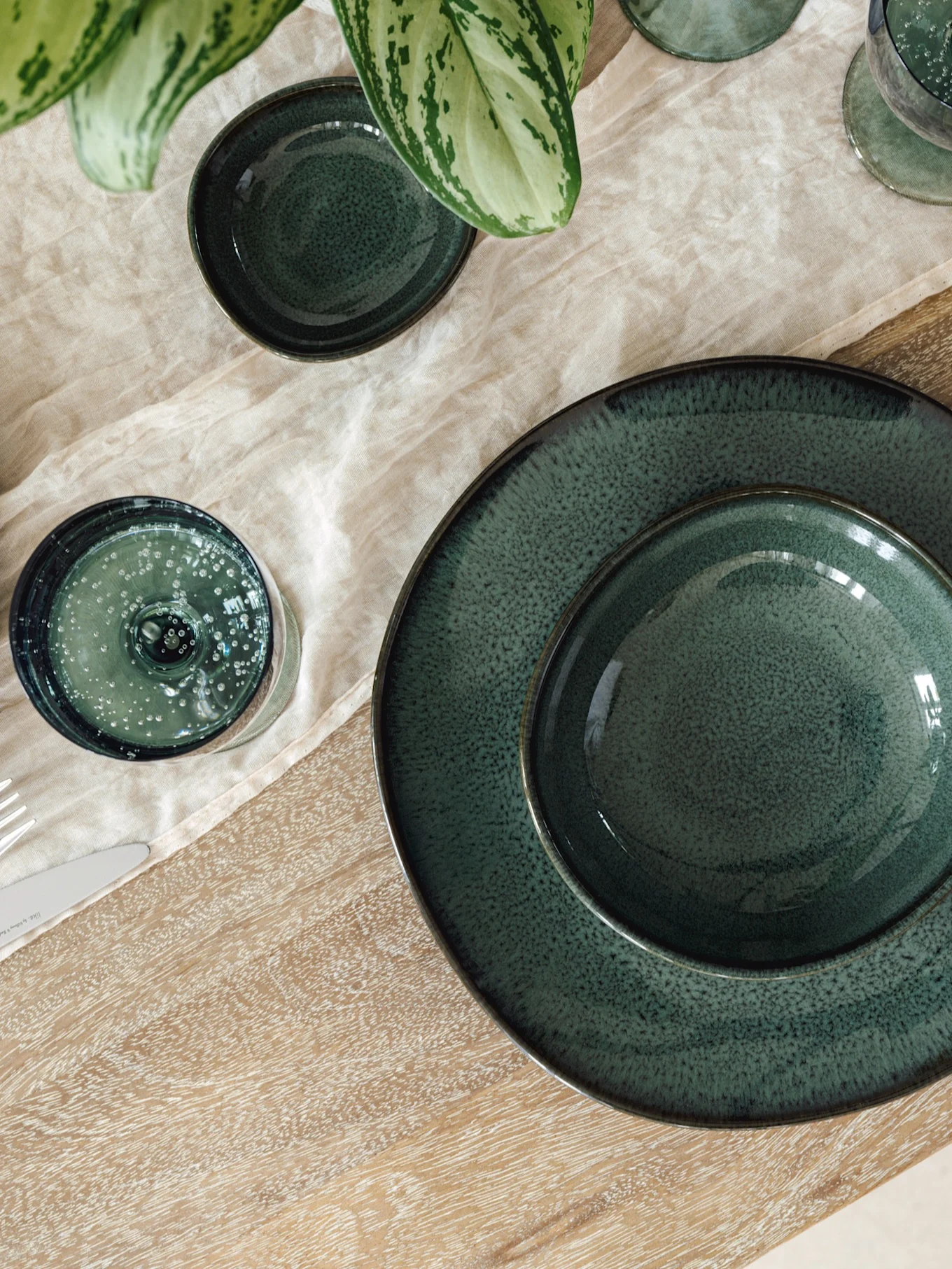 Table dressée avec assiettes, bols et verres en céramique vert foncé sur nappe en lin clair et table en bois, avec plante verte.