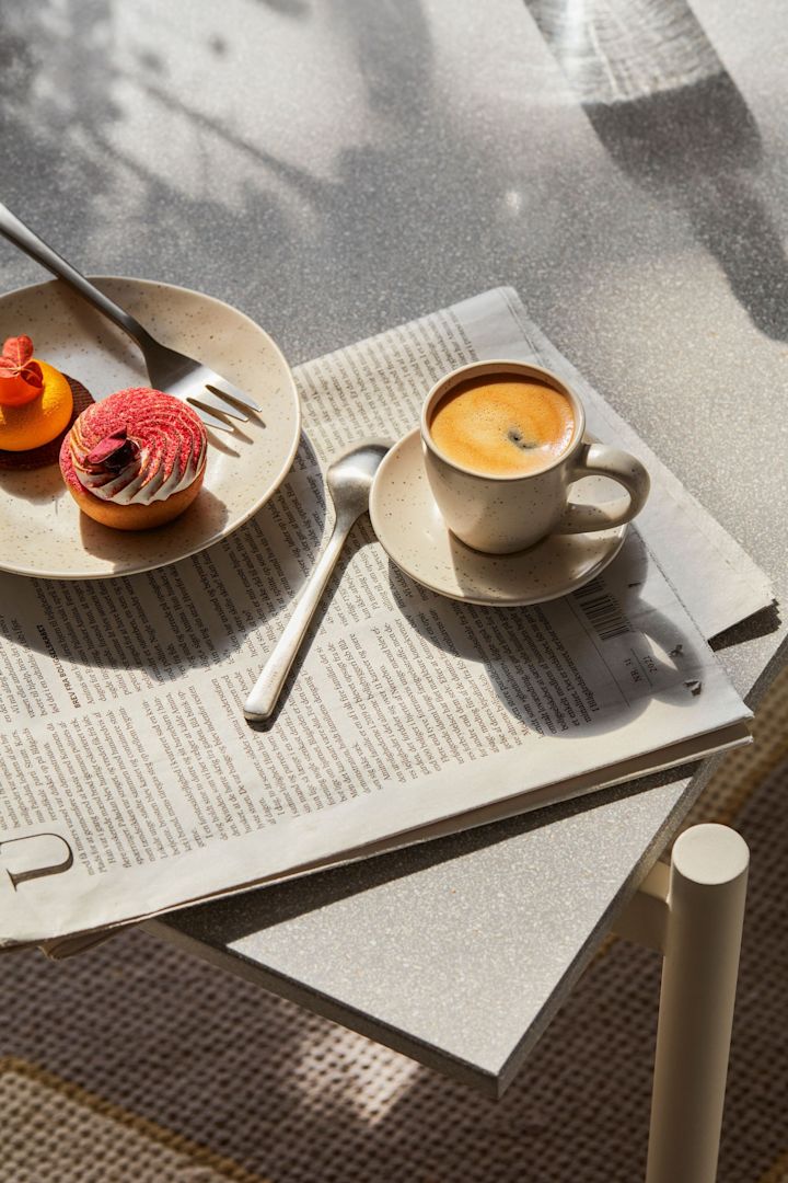 La tasse à espresso Nordic Vanilla de Broste Copenhagen sur une table de petit-déjeuner, posée sur un journal quotidien, avec une assiette de gâteau à côté.