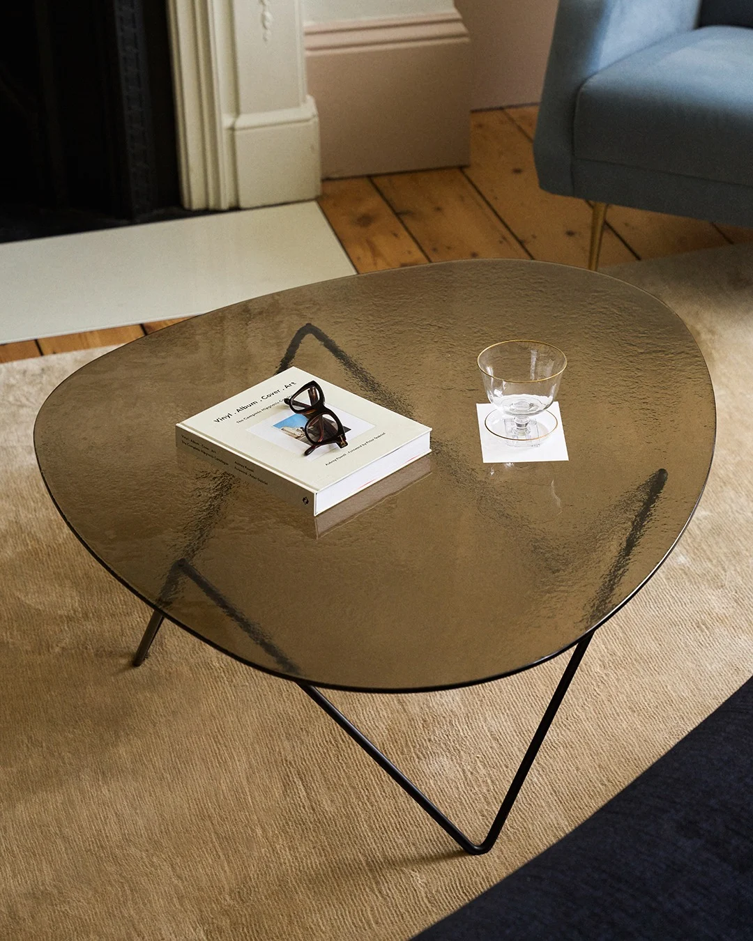Table basse en verre foncé de forme organique, avec un livre, des lunettes écaille de tortue et un verre à bord doré sur un sous-verre.