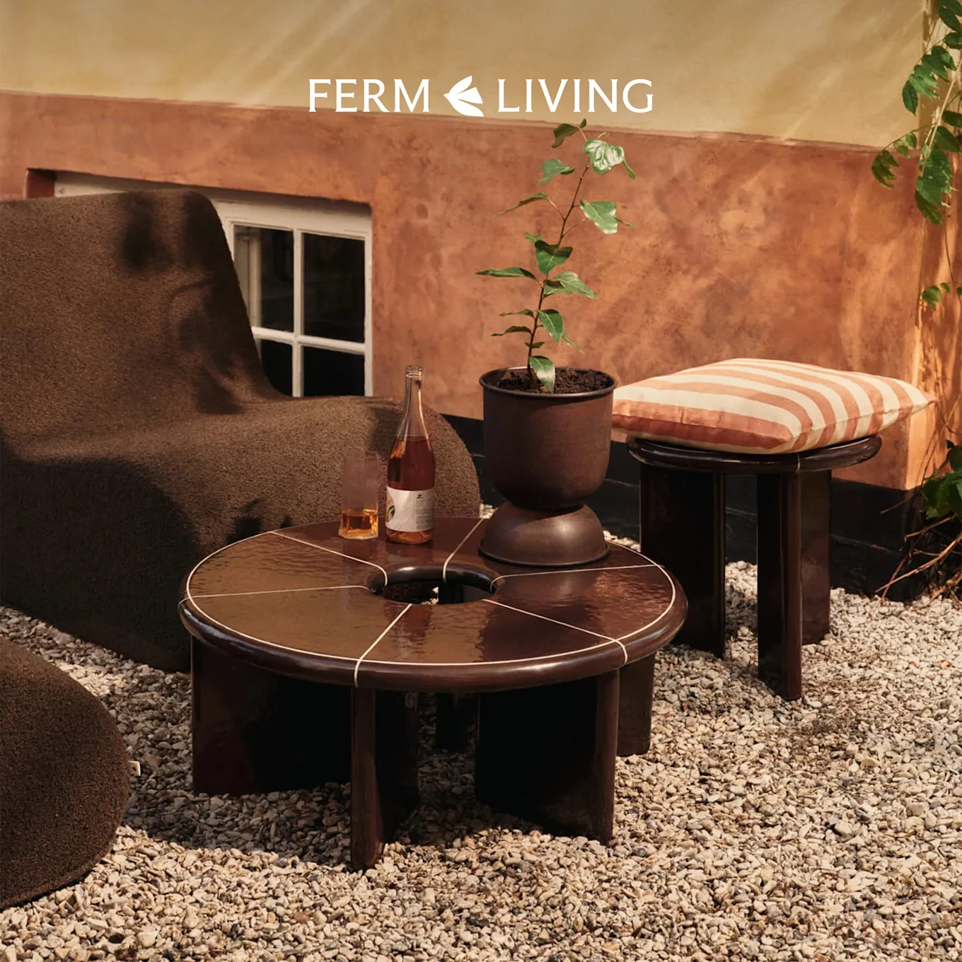 Fauteuil marron, table ronde avec boissons, plante, coussin rayé sur tabouret sur patio de gravier.