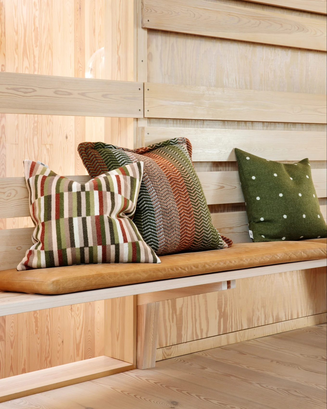 Trois coussins décoratifs avec différents motifs sur un coussin de banc en cuir marron clair, devant un mur en bois.