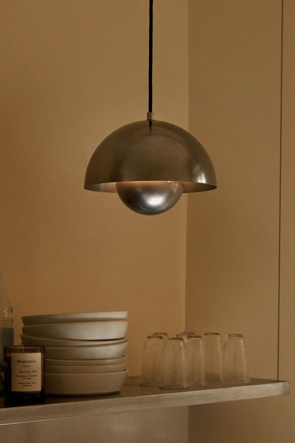 Une suspension métallique en forme de dôme est suspendue au-dessus d'une étagère avec des bols, des verres, des bouteilles et une bougie sur un mur marron chaud.