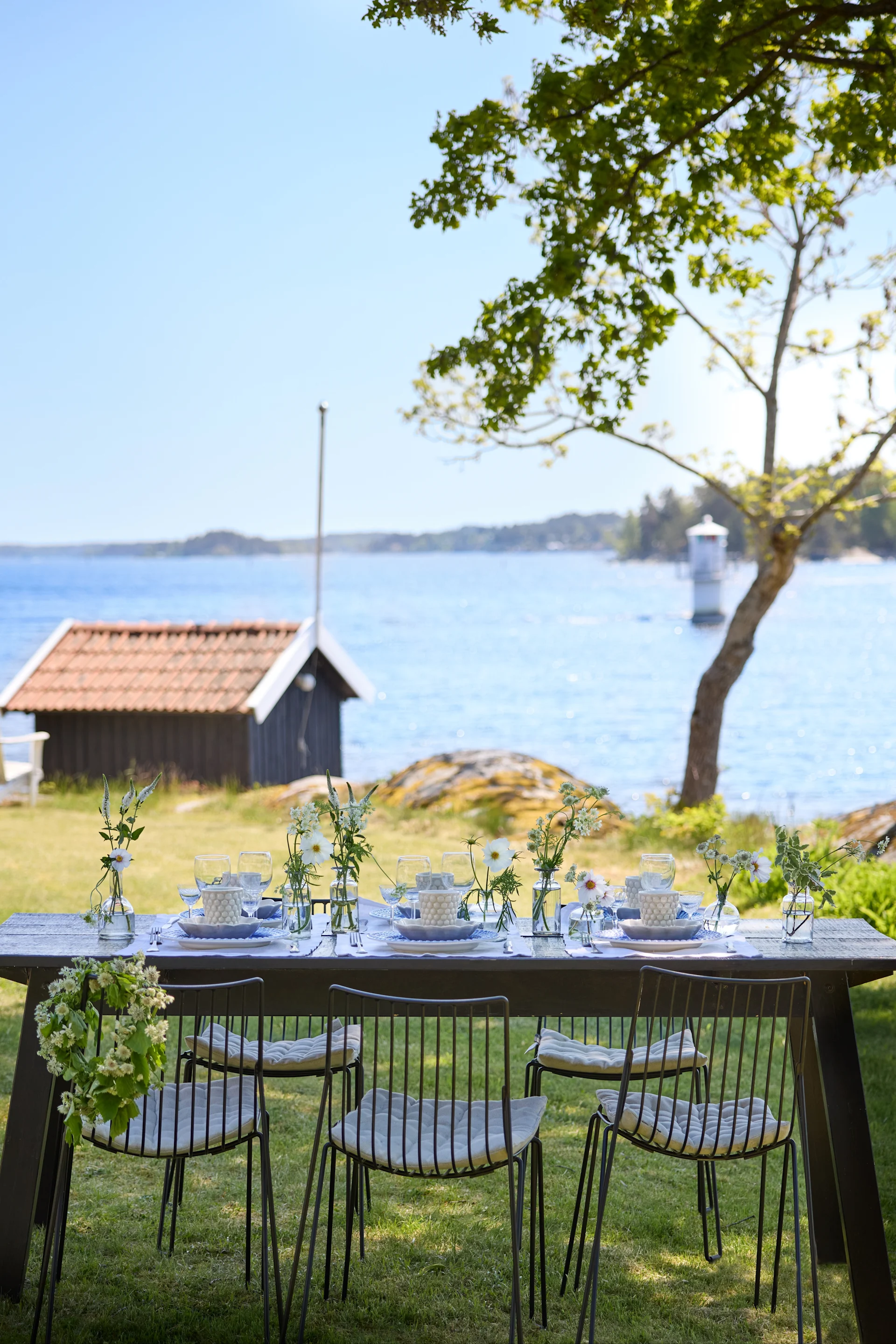 Une table dressée avec de la vaisselle Mateus en bleu et blanc, dans un décor verdoyant avec vue sur une petite maison et un lac.