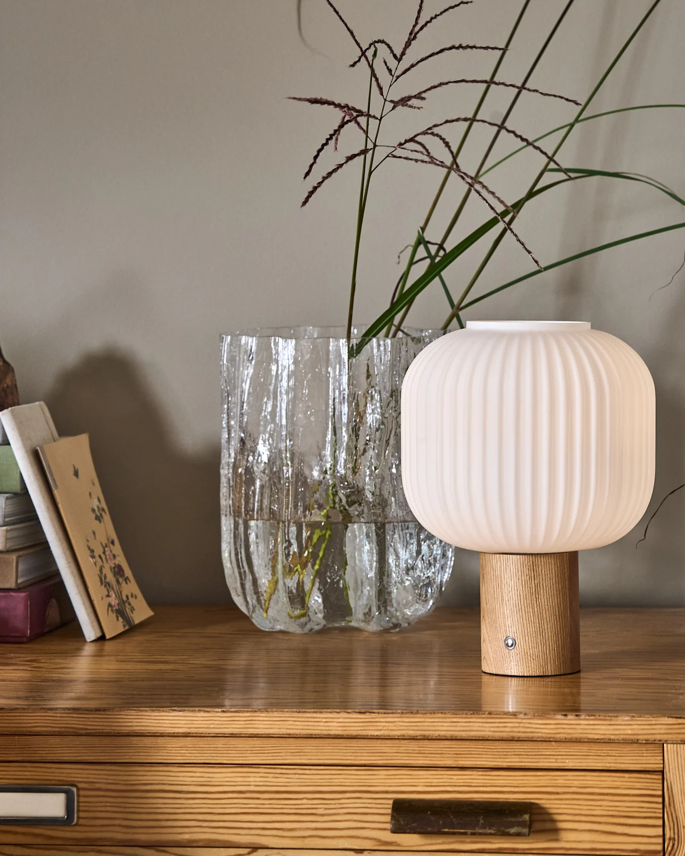 Une lampe côtelée en forme de champignon avec une base en bois à côté d'un vase en verre texturé avec des plantes et une pile de livres sur une commode en bois.