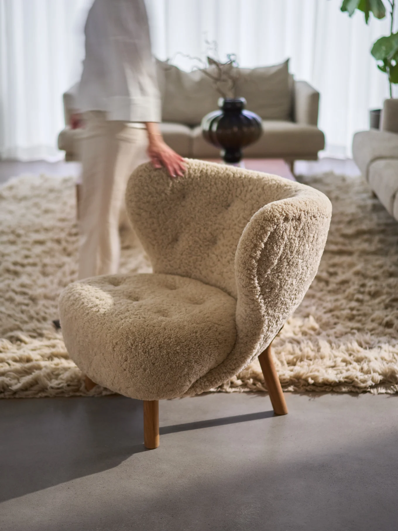 La main d'une personne repose sur un fauteuil beige douillet et texturé avec des pieds en bois, sur un tapis shaggy dans un salon.