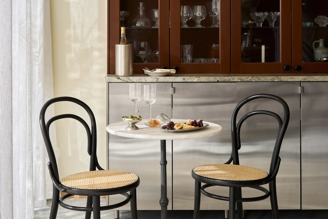 Coin repas confortable avec table en marbre, deux chaises noires, plateau de fromages, fruits, olives et verres à vin.