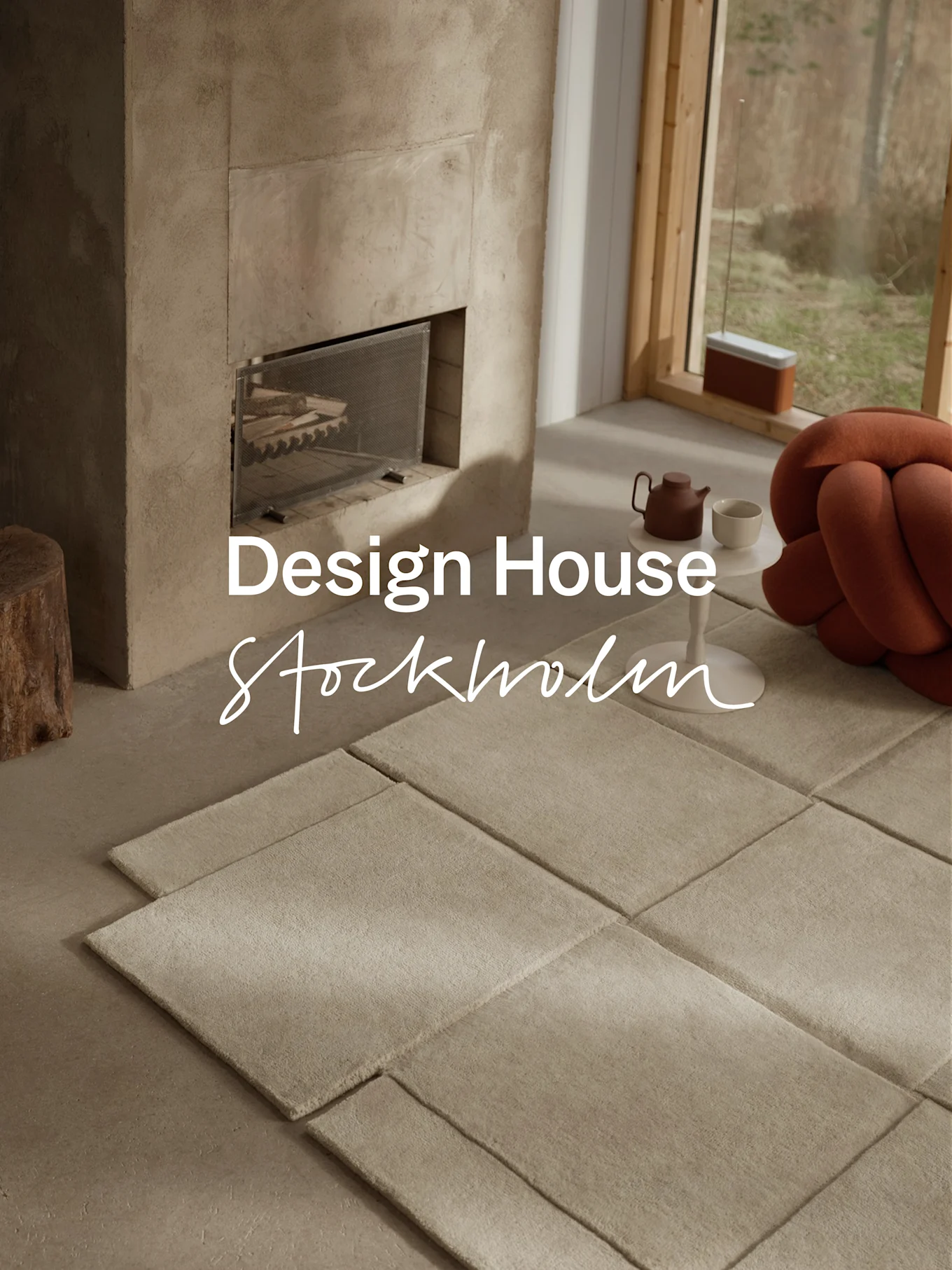 Salon moderne avec cheminée en béton, tapis modulaires de couleur claire, une théière et un coussin noué orange près d'une grande fenêtre.