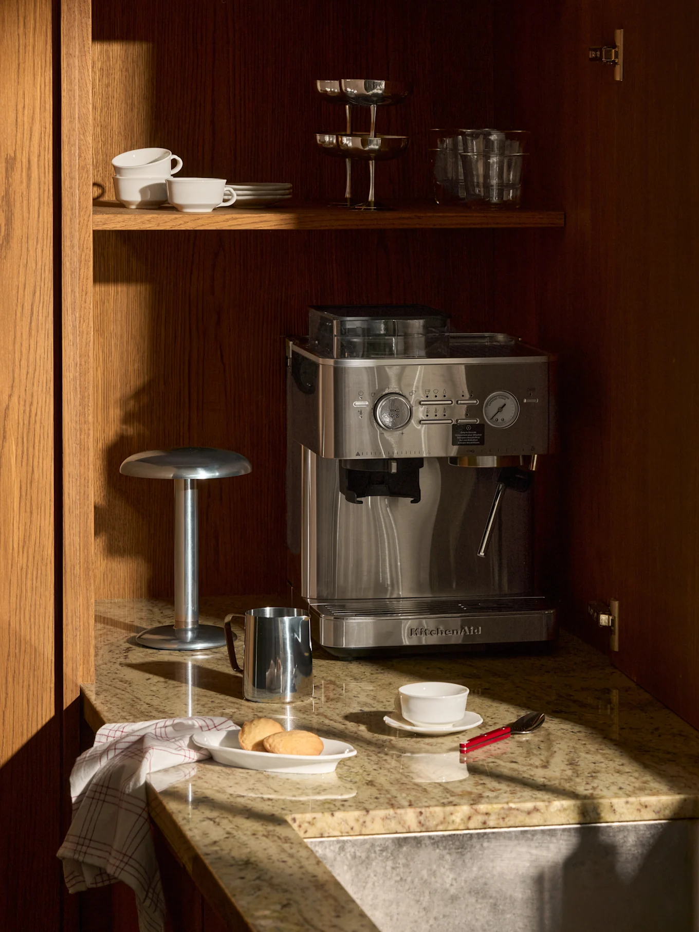 Machine à expresso en inox sur comptoir granit, dans armoire bois. Dessus, tasses, verres. Sur comptoir, biscuits, tasse, pot à lait.