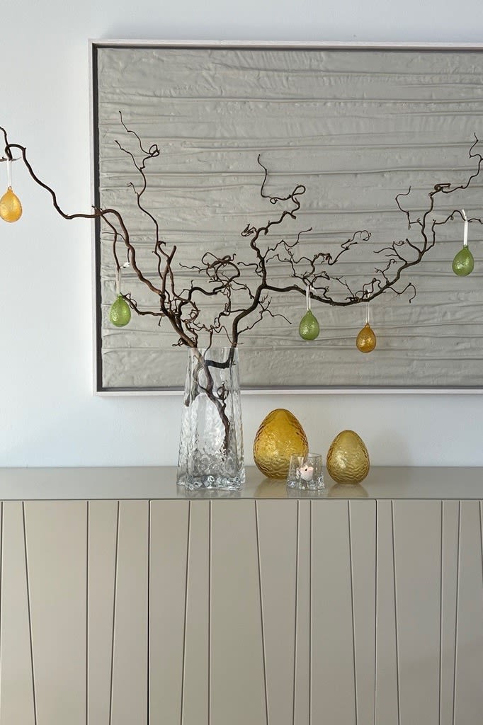 Un arbuste de Pâques est posé sur une commode dans un vase Gry, décoré de pendentifs de Pâques Gry, à côté d'œufs en verre Gry, le tout de Cooee Design.