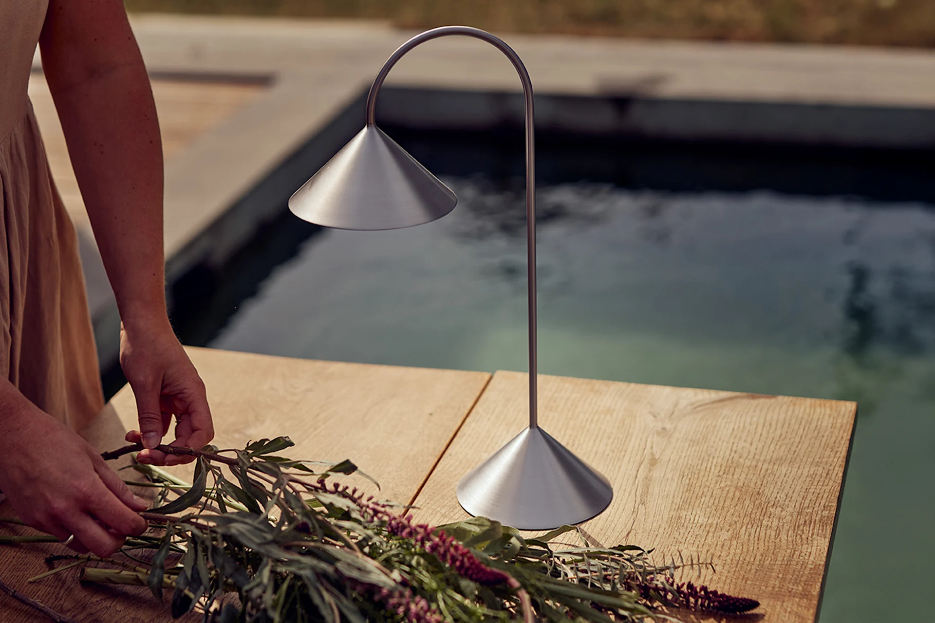 Mains arrangeant des fleurs sur une table en bois à côté d'une lampe de table en métal brossé près d'une piscine.