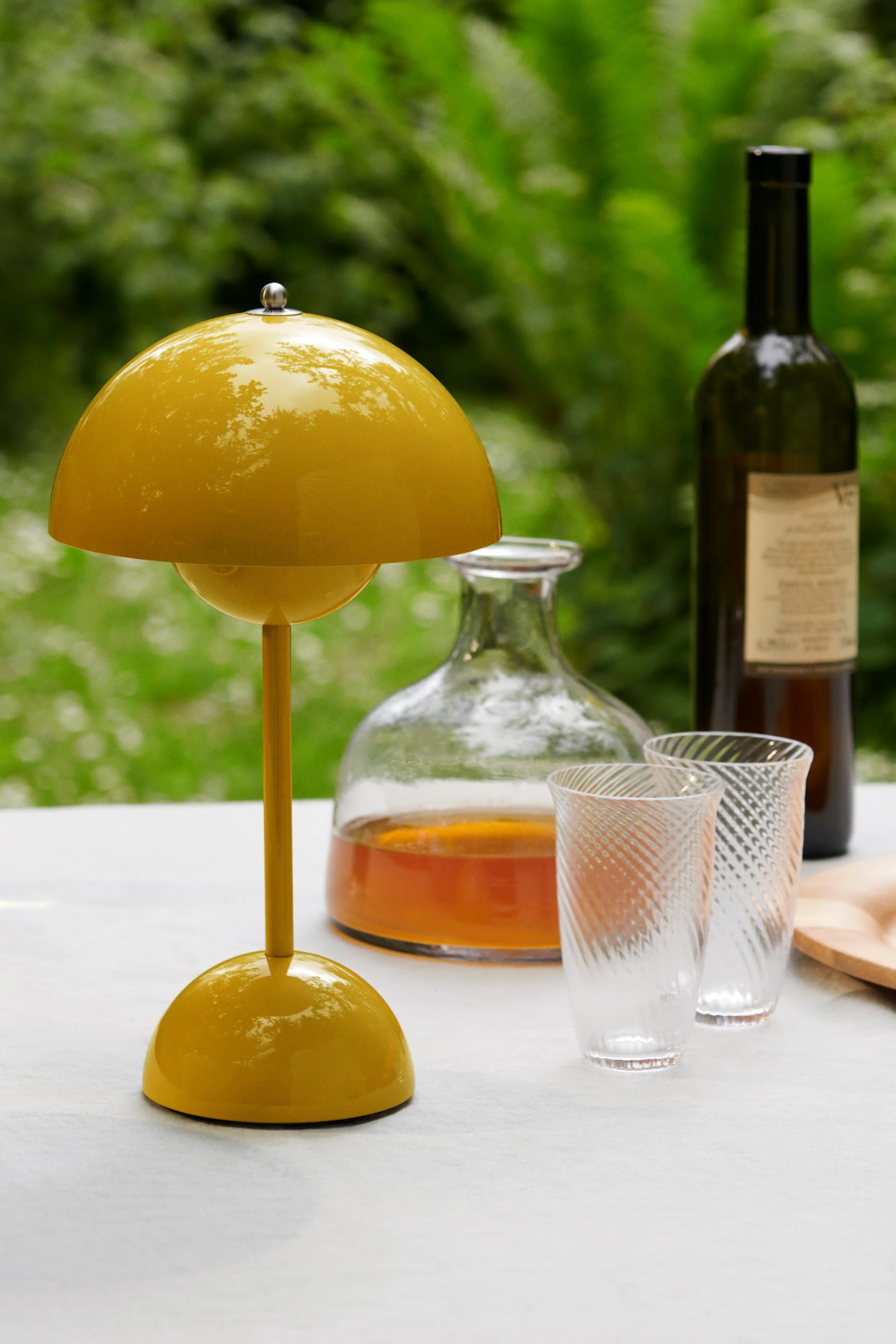 Une lampe de table jaune vif en forme de champignon sur une table blanche, avec une carafe, deux verres à motifs et une bouteille de vin derrière, sur fond vert.