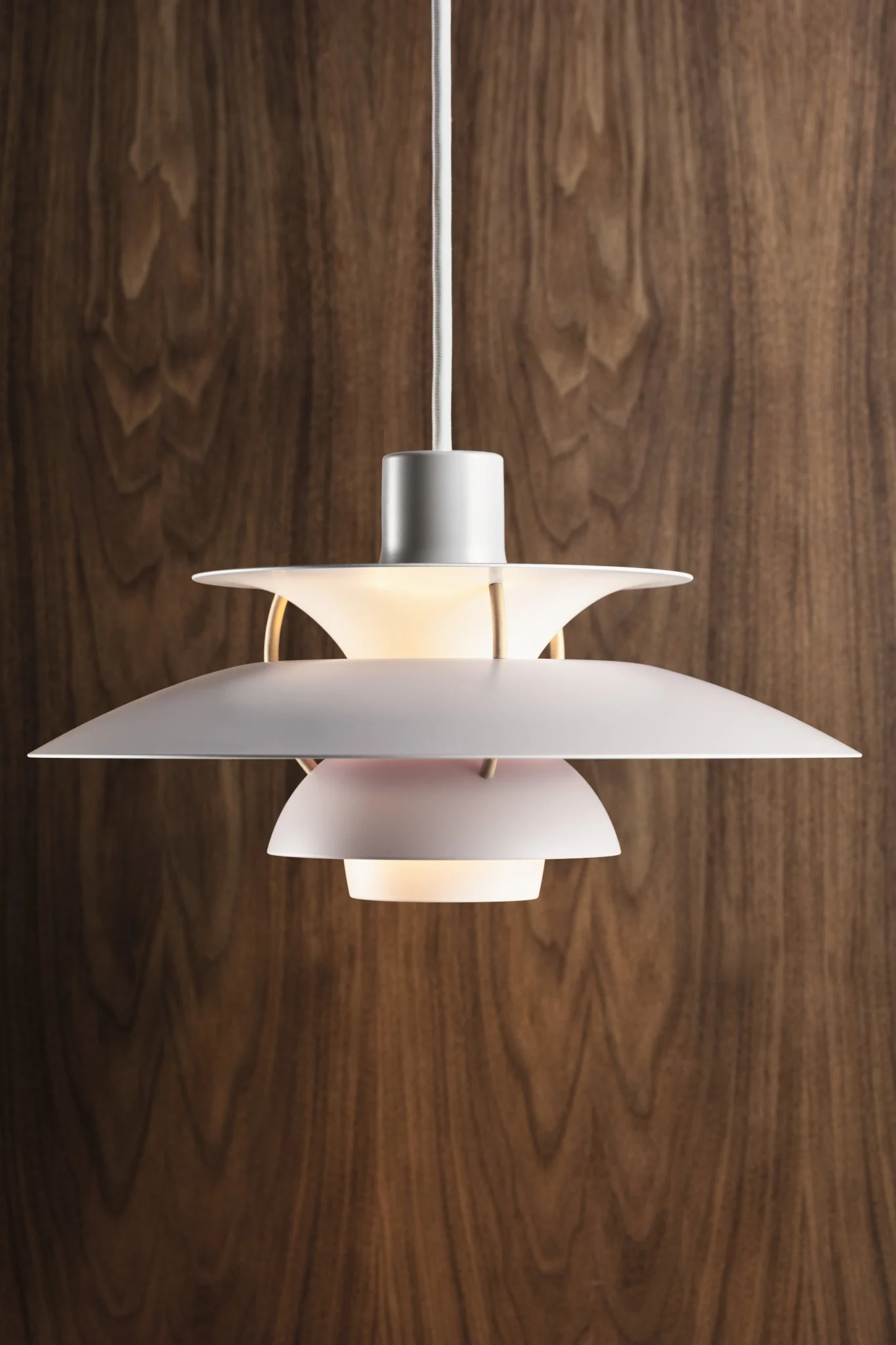 Une lampe suspension blanche à plusieurs niveaux avec une lueur chaude, suspendue contre un mur en panneaux de bois foncé.