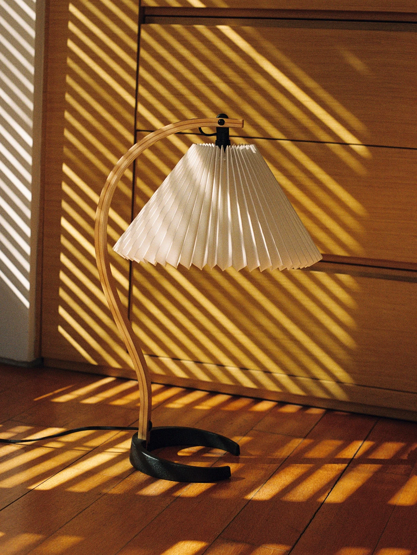 Lampe de bureau avec un pied en bois courbé et un abat-jour plissé, éclairée par des rayures de soleil diagonales sur un parquet.