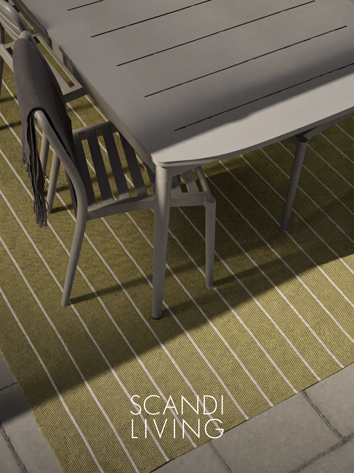 Vue plongeante d'une table et chaise de salle à manger grises d'extérieur avec un plaid foncé sur un tapis rayé vert et blanc.