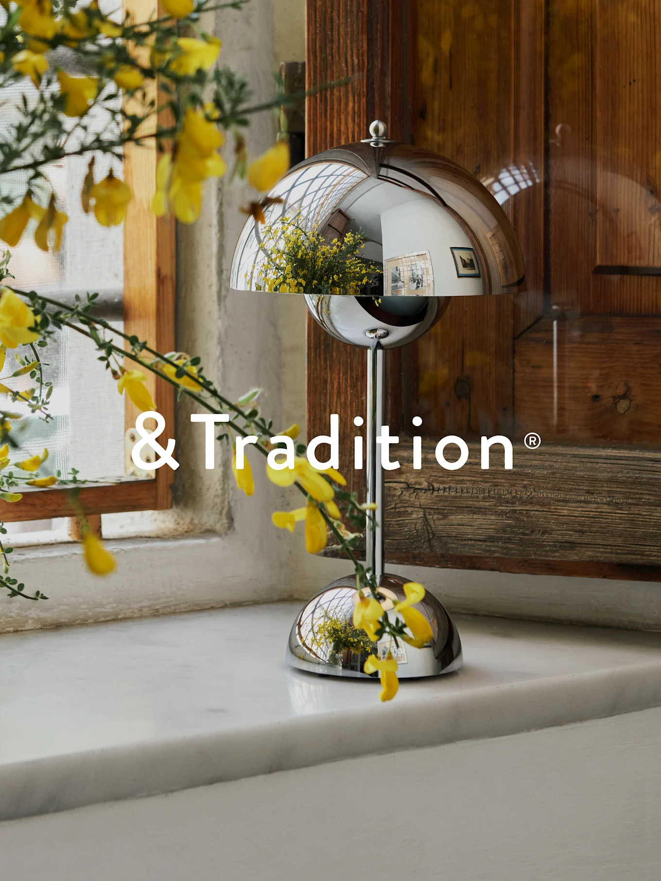 Une lampe de table argentée en forme de dôme sur un rebord de fenêtre blanc, avec des fleurs jaunes au premier plan. La lampe reflète la fenêtre et les fleurs. Logo '&Tradition'.