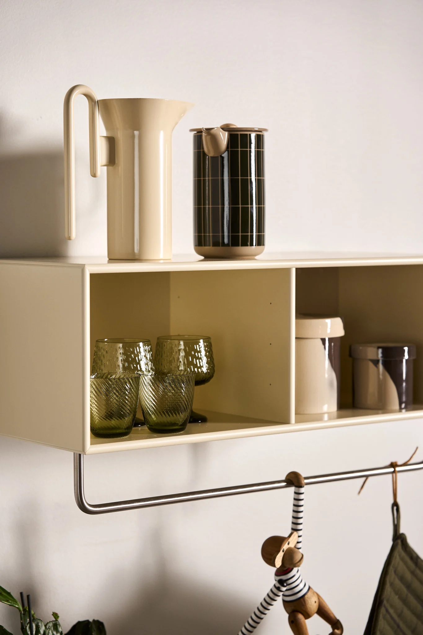 Une étagère murale beige clair contient une carafe crème, un récipient noir, des verres verts texturés et des pots à motifs. Un singe en bois pend en dessous.