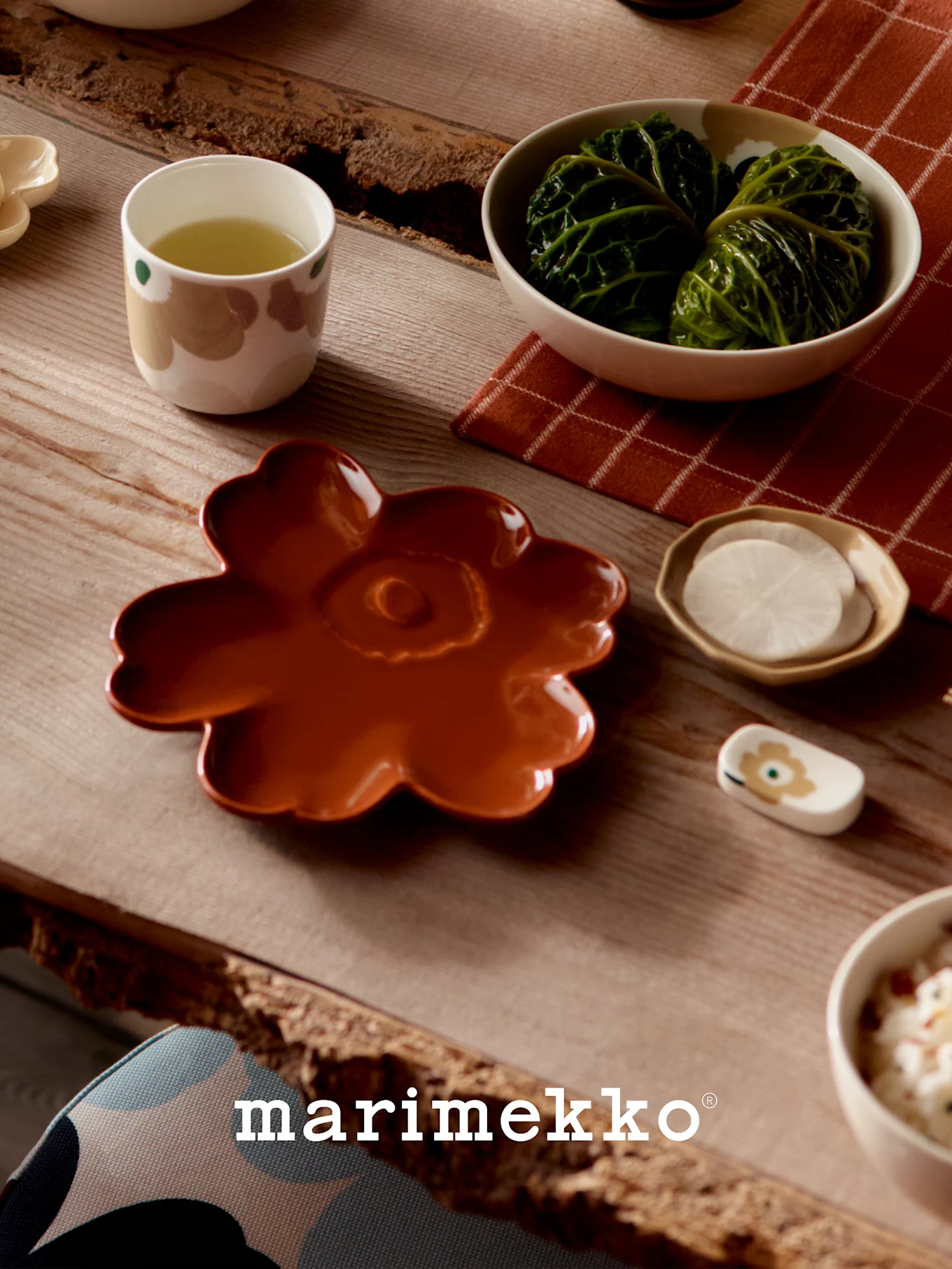 Table dressée avec de la vaisselle Marimekko : une assiette florale couleur rouille, une tasse de thé vert, des roulés de chou et des radis tranchés.