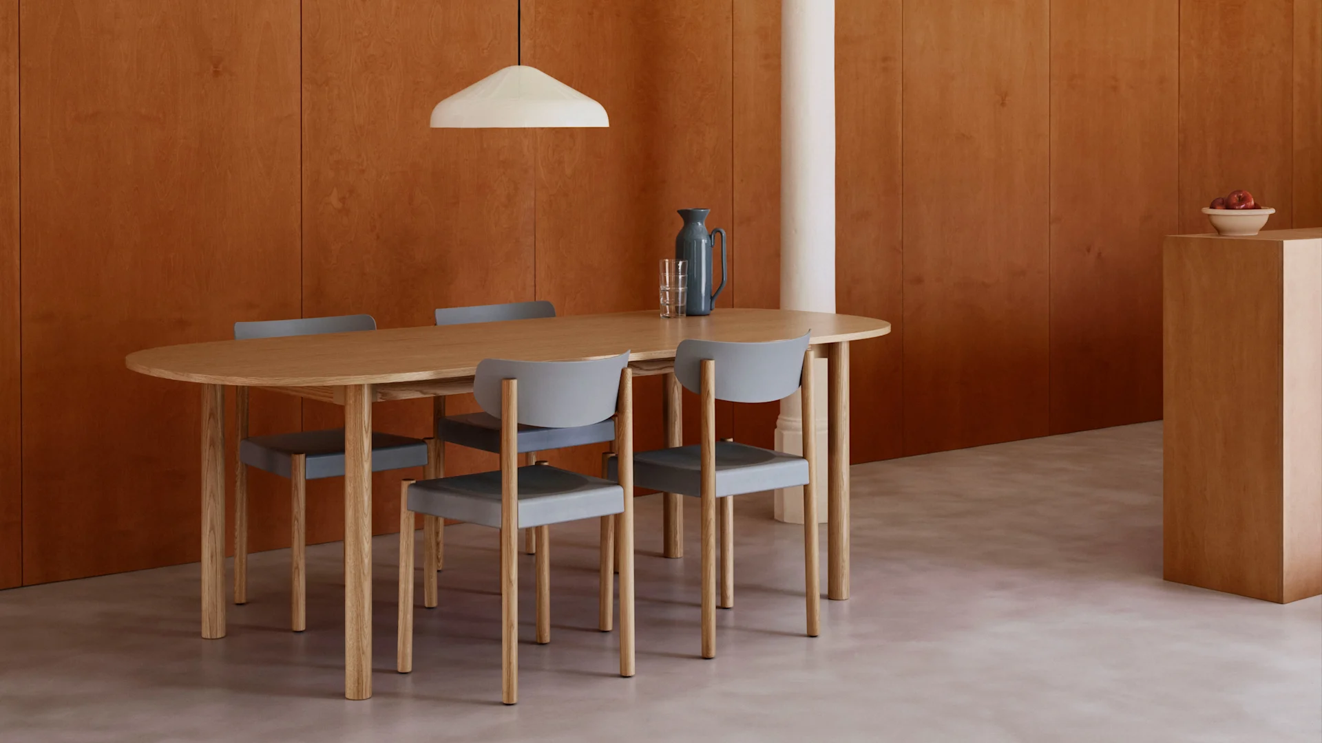 Salle à manger moderne avec table ovale en bois, quatre chaises grises, murs lambrissés chauds, sol clair et suspension blanche.