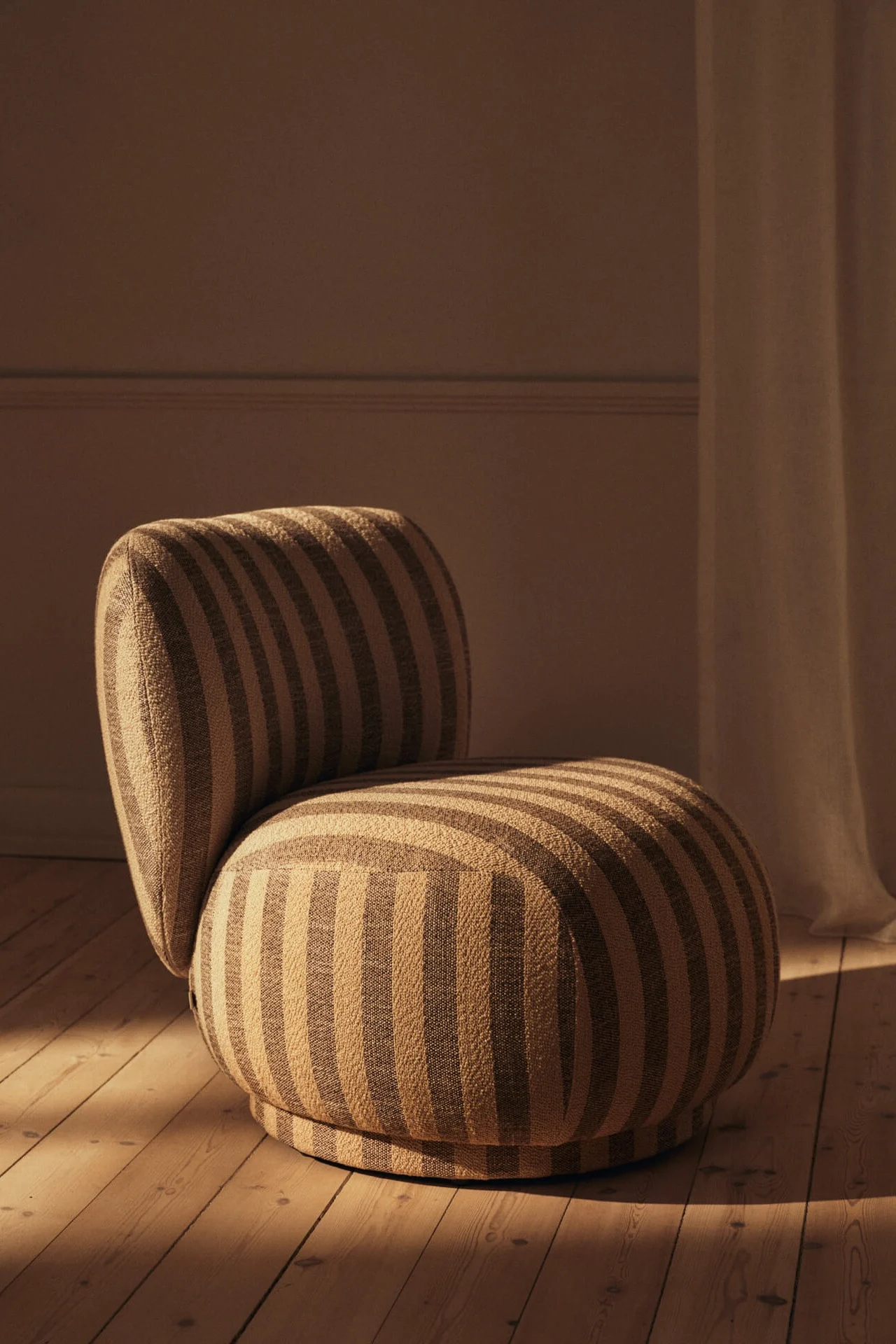 Fauteuil moderne et arrondi à rayures beiges et marron sur un parquet, baigné d'une lumière chaude.