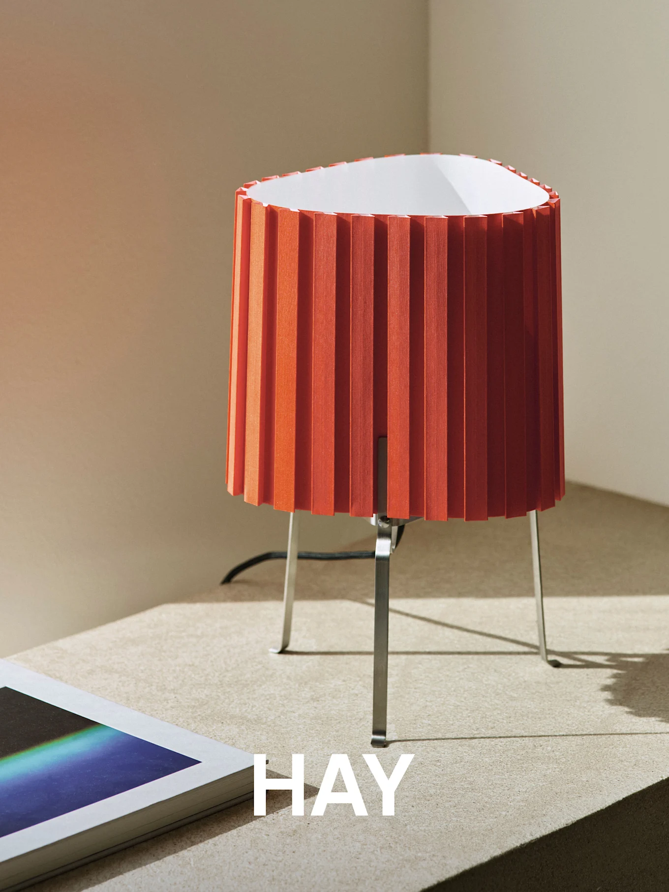 Lampe de table plissée orange avec une base en métal sur une surface texturée, à côté d'un livre. Le mot 'HAY' est superposé.
