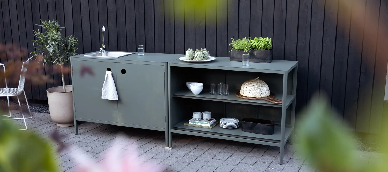 Un module de cuisine d'extérieur gris-vert, avec un évier et un essuie-mains à gauche, ainsi que des étagères sur lesquelles sont disposés des ustensiles de cuisine et des pots d'herbes aromatiques à droite.